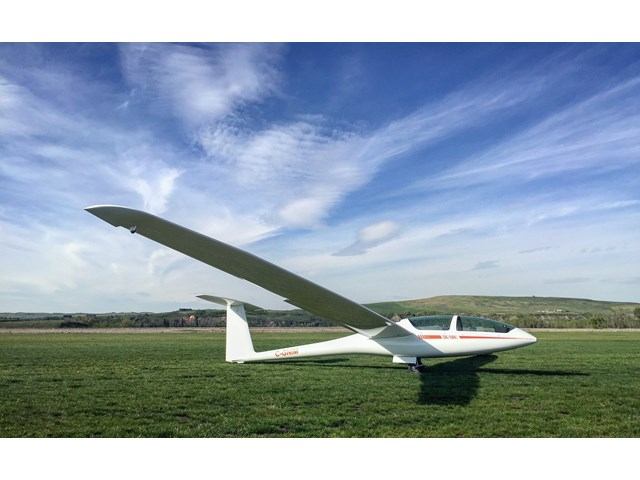 White glider on a grassy airfield under a bright blue sky with wispy clouds.