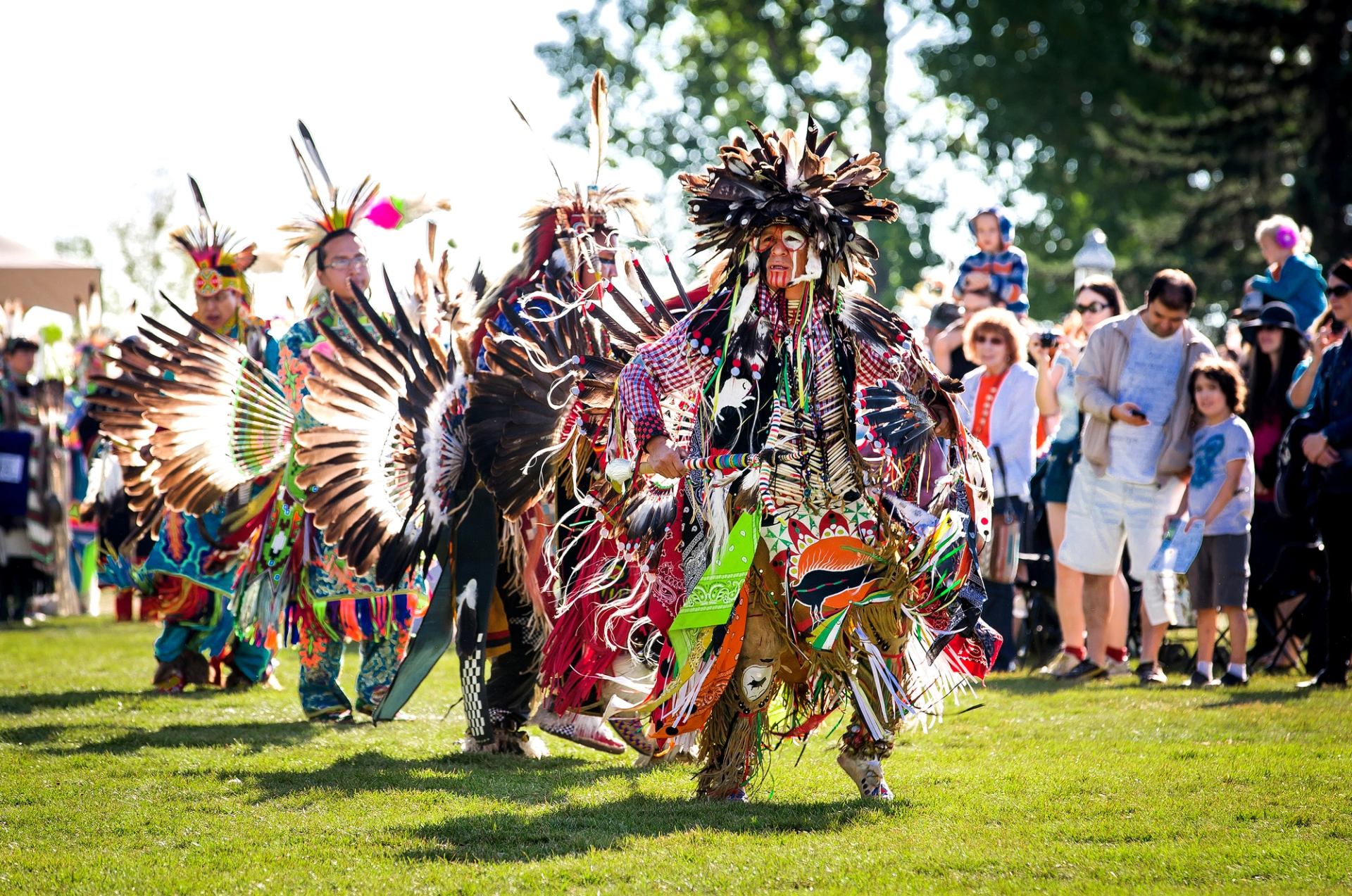 Powwow dancers in colourful feathered regalia perform on grass before a watching crowd.