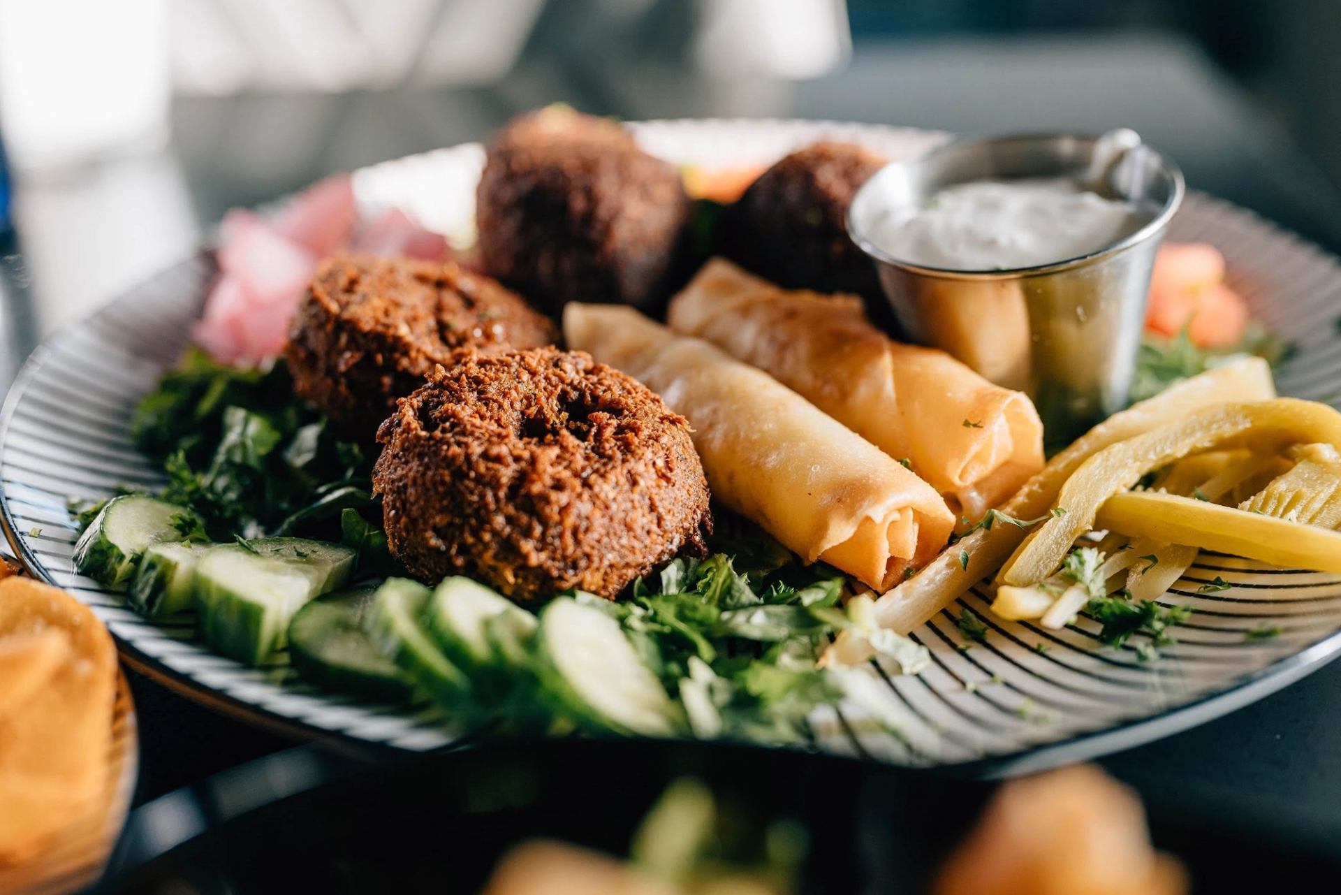 A platter features falafel, vegetable rolls, fresh vegetables, and creamy dipping sauce.