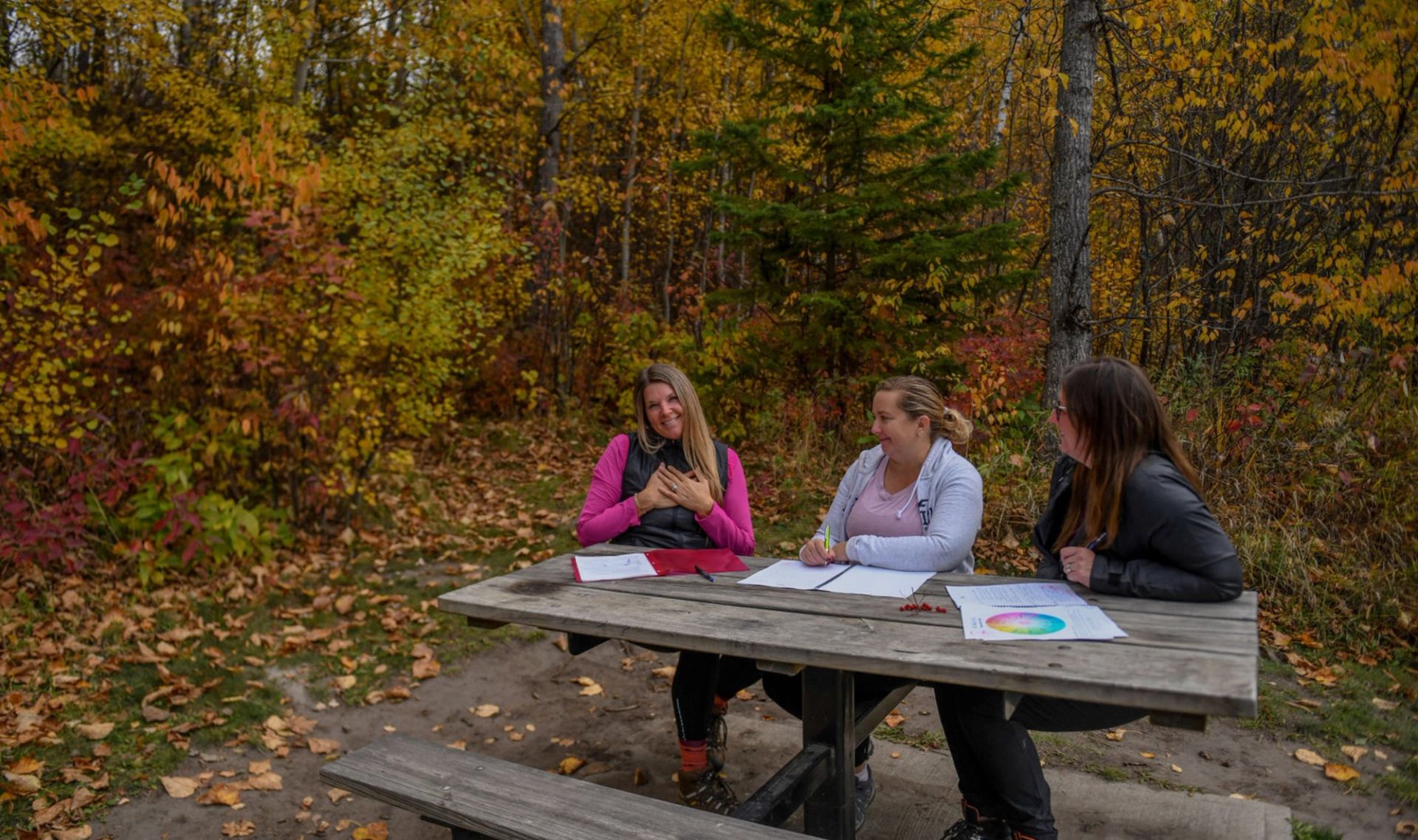 A small group seated at a picnic table in a forest clearing with autumn colors.