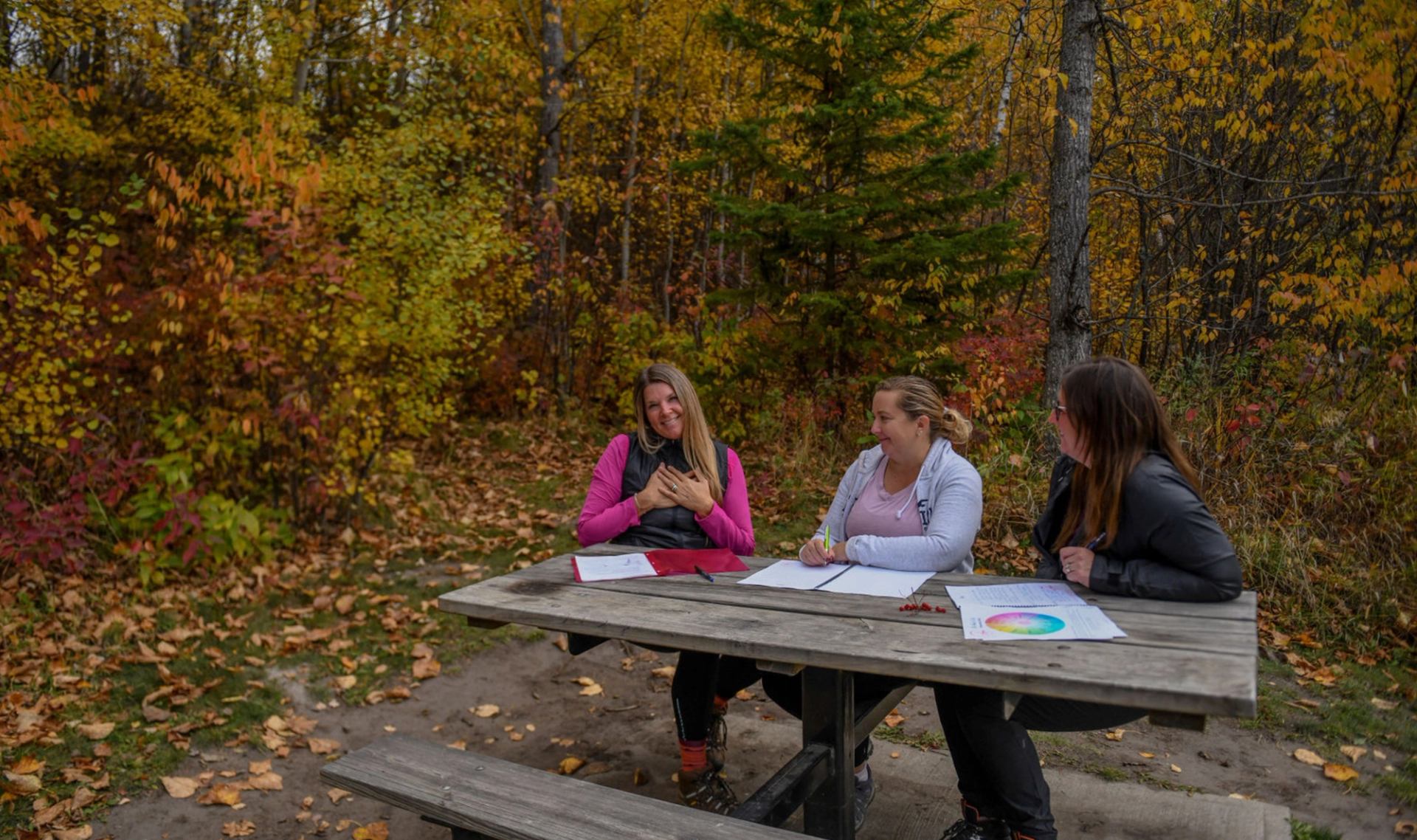 A small group seated at a picnic table in a forest clearing with autumn colors.