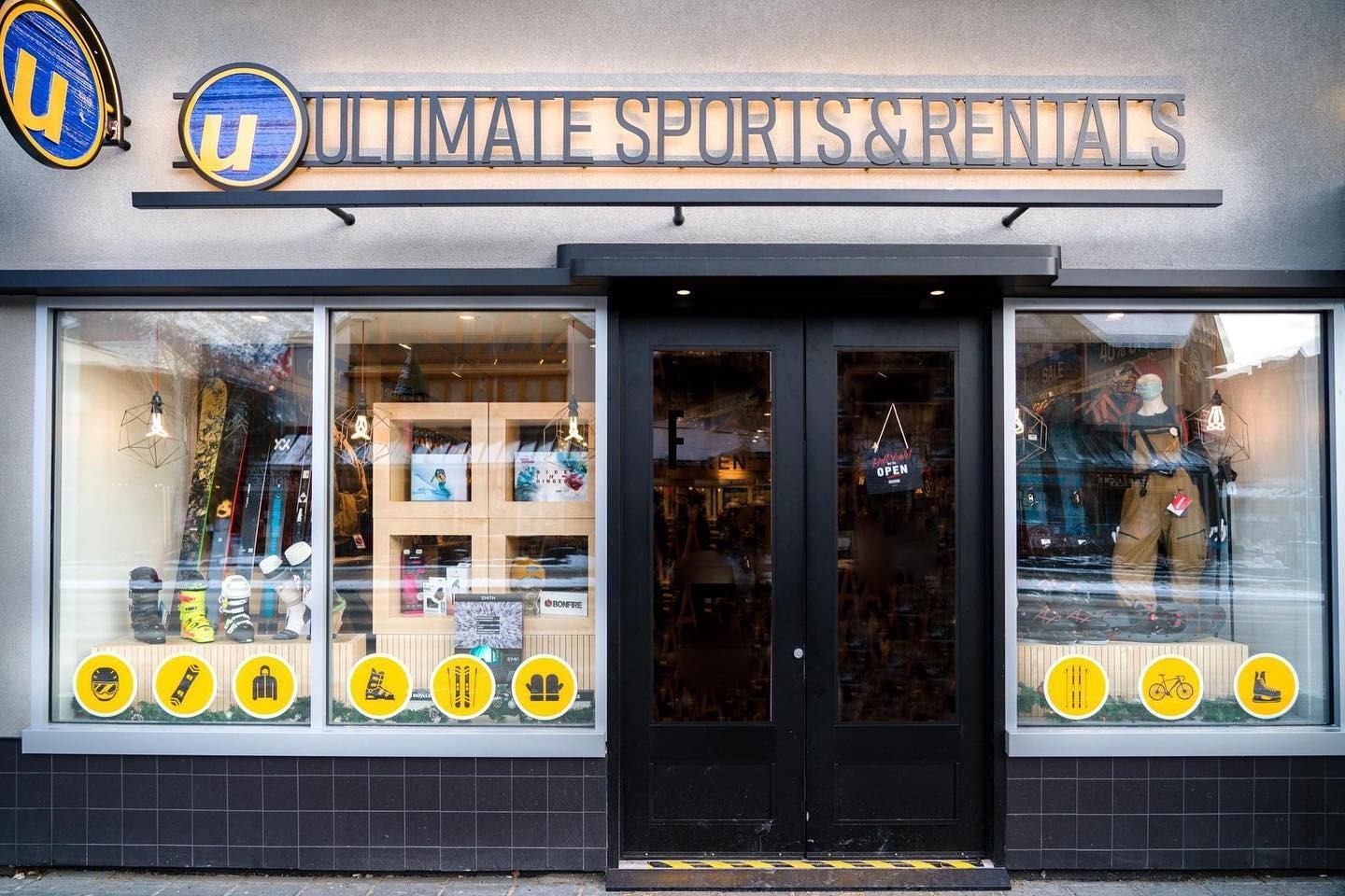 Storefront of "Ultimate Sports & Rentals" with large windows displaying sports equipment. Bright yellow signage and black double doors convey a modern feel.