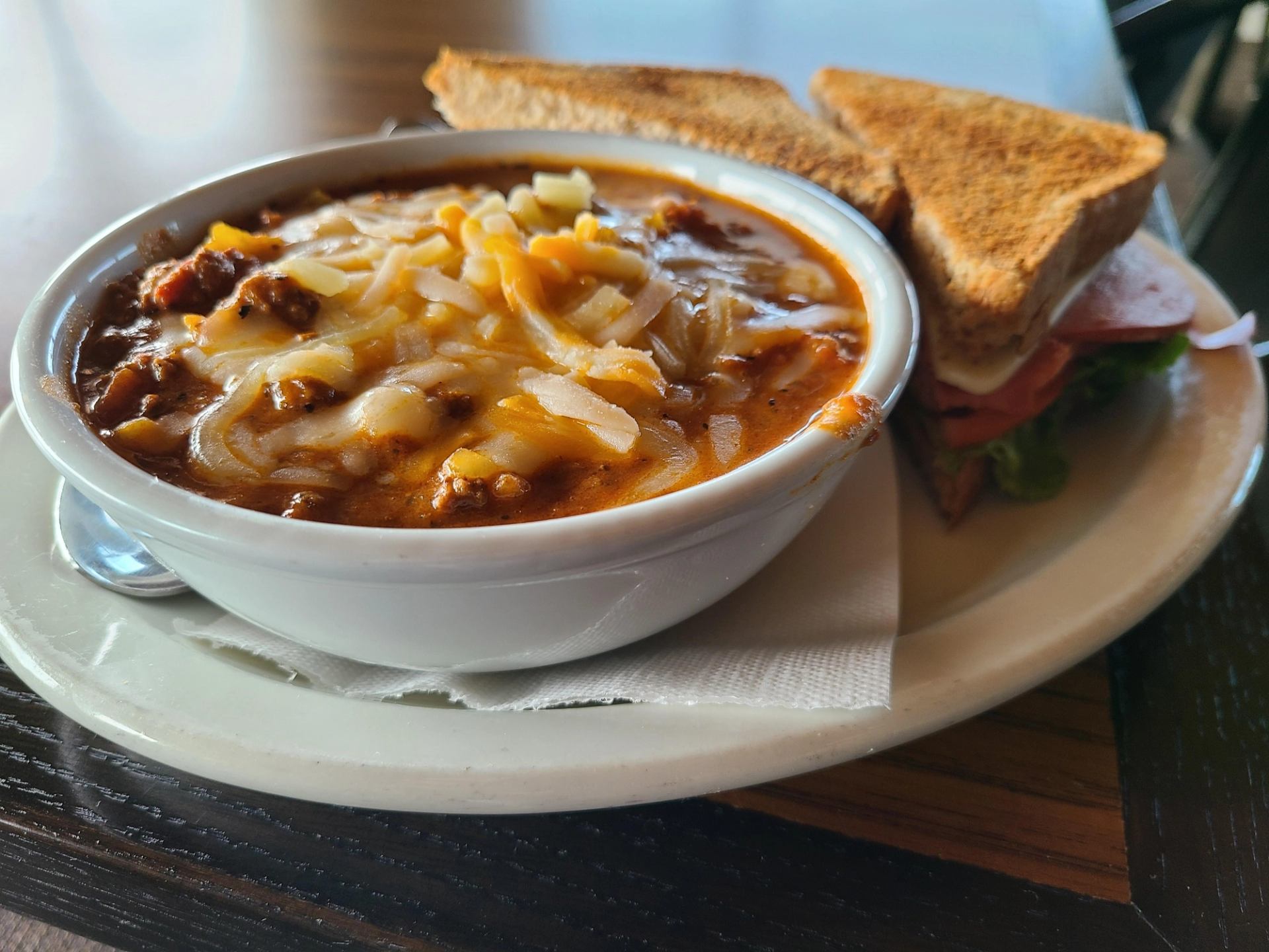 A bowl of beef chili topped with melted cheese served alongside a toasted deli sandwich.