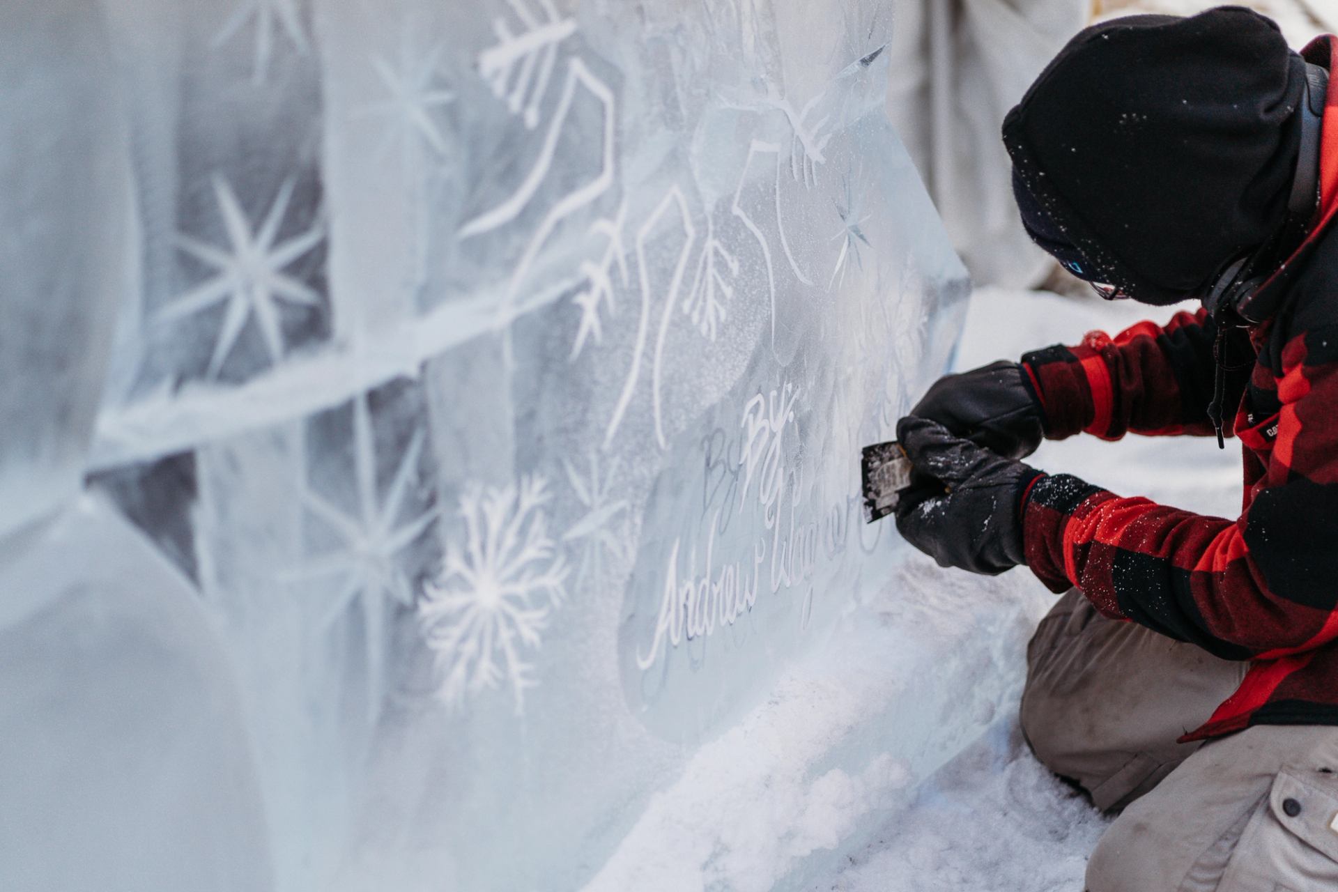 Person carving intricate snowflake and animal designs into a large ice block at an outdoor winter event.