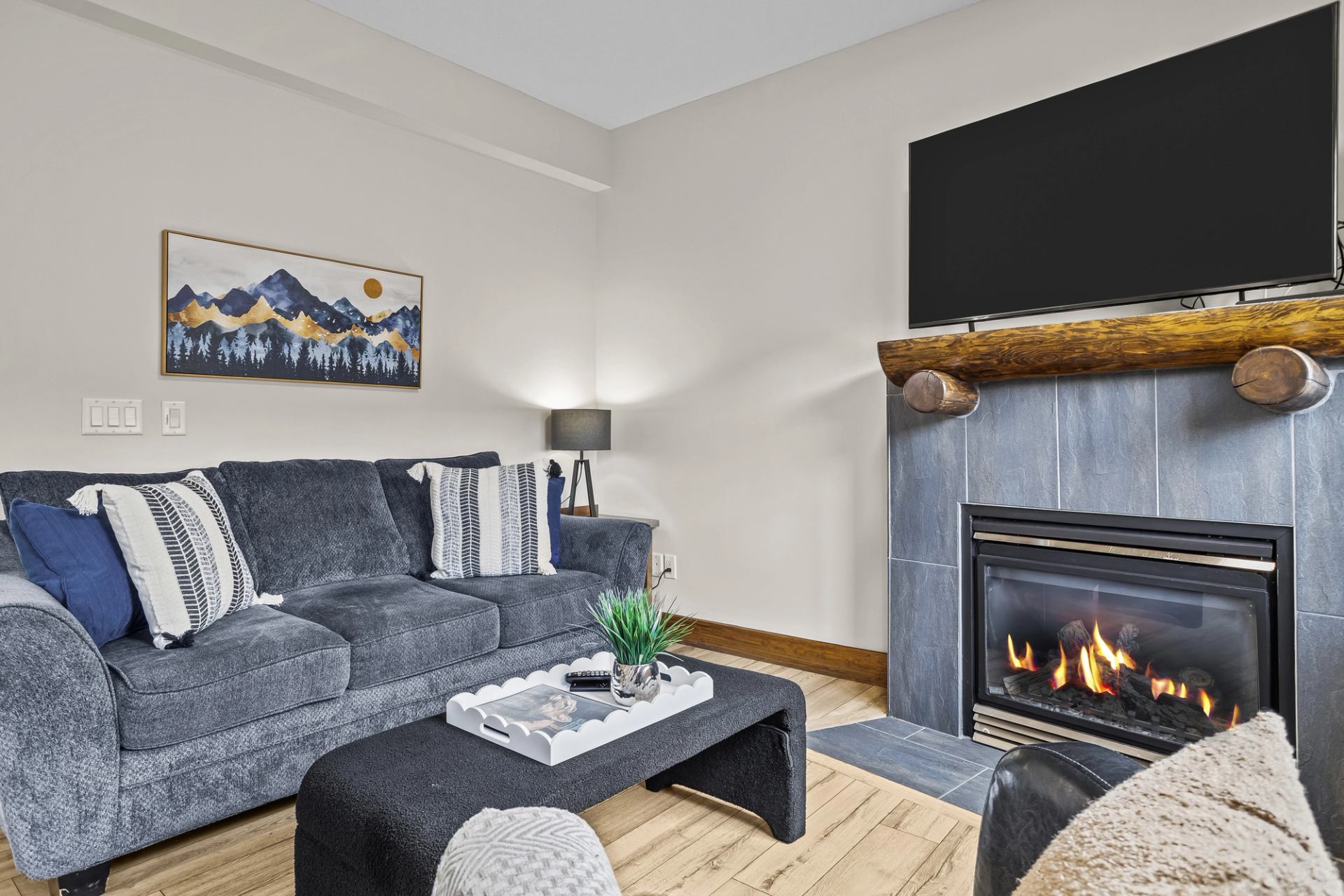 Living room with grey sofa, lit fireplace, wall-mounted television and mountain artwork.
