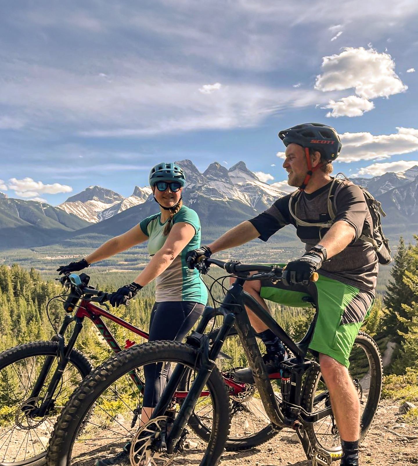 Two cyclists with helmets on a trail overlooking rugged mountain peaks
