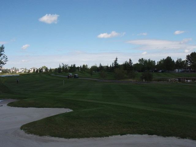Golf course fairway with sand bunker, trees, and houses under a blue sky.