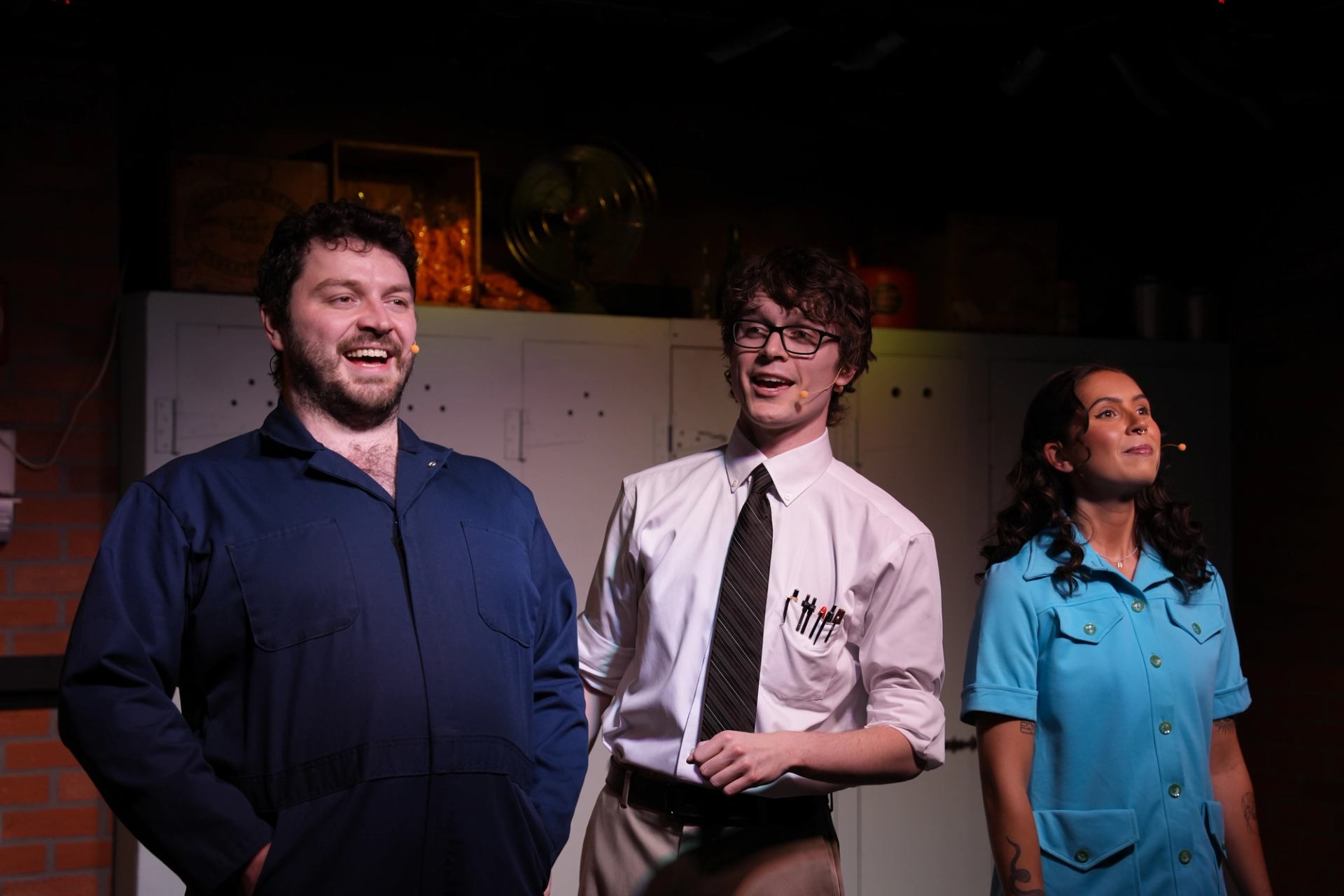 Three performers on stage wearing microphones and costumes, standing before lockers and props.