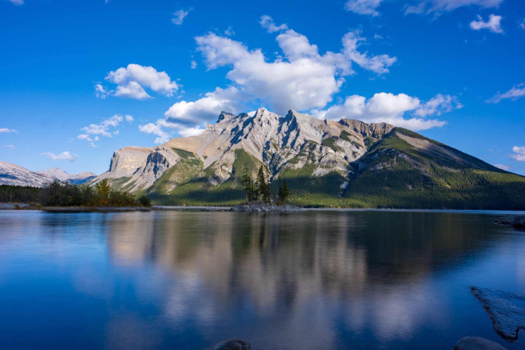 Mountain range reflected in a calm lake under a bright blue sky with scattered clouds.