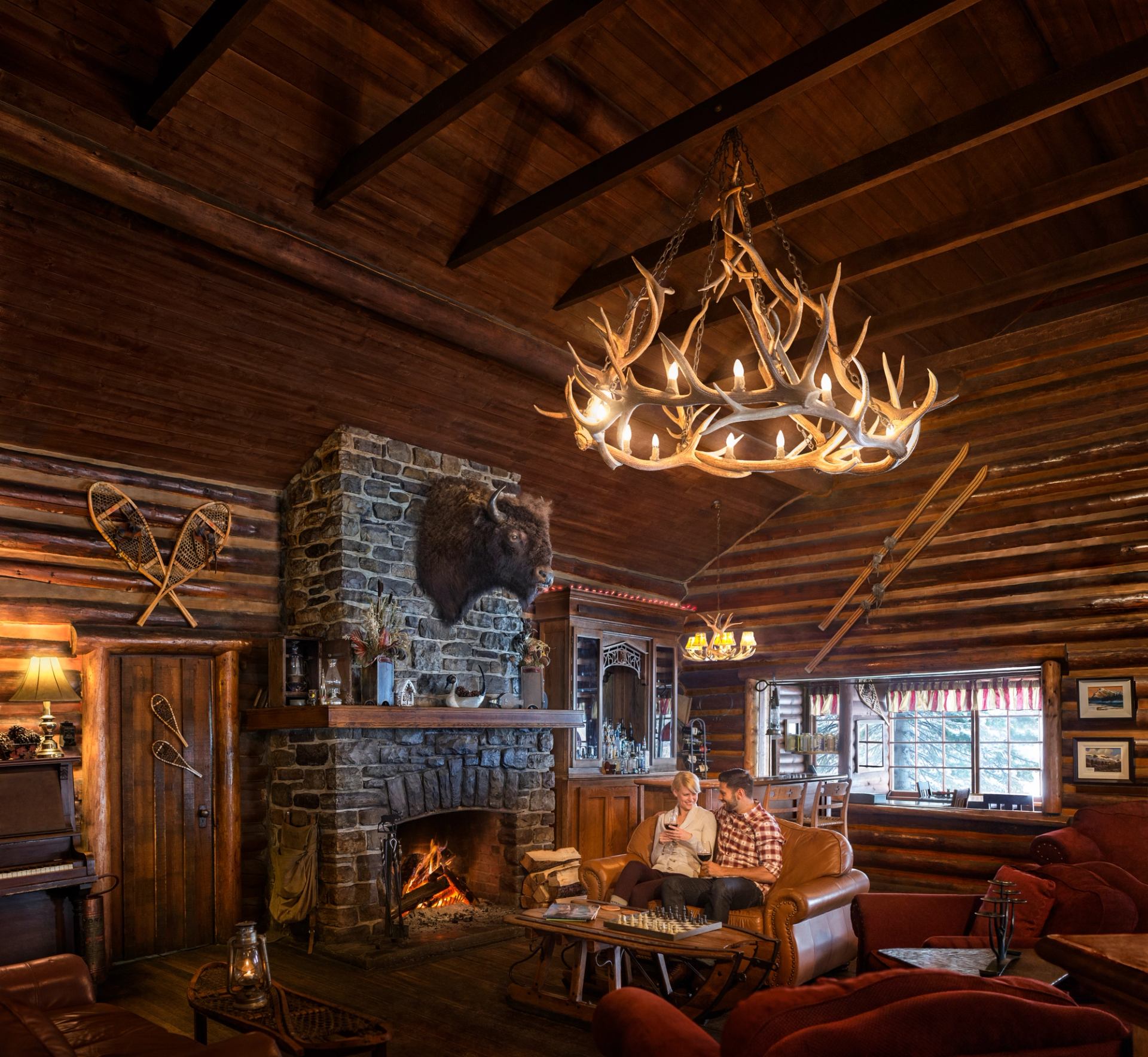 Two people chat by a fireplace in a rustic log cabin interior drinking wine.