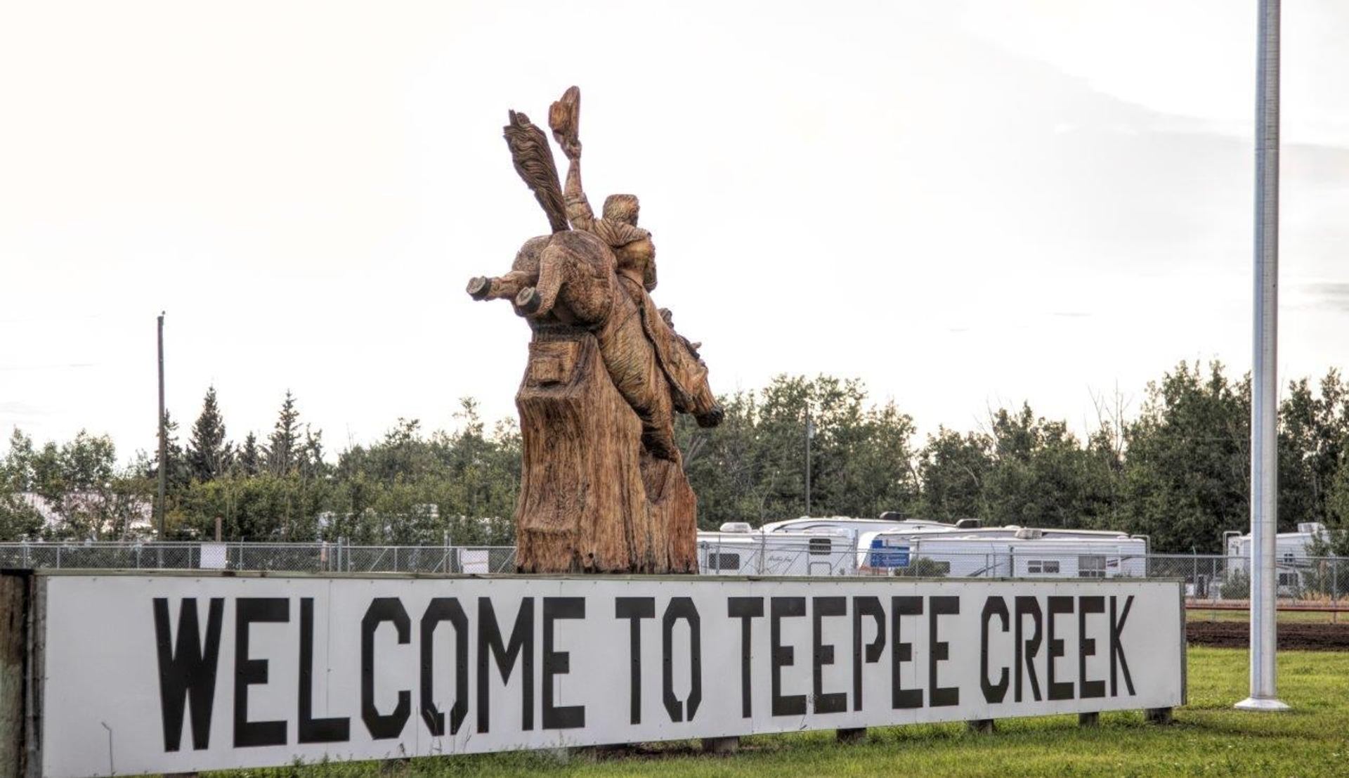 Wooden rider statue above large Welcome to Teepee Creek sign near trees and trailers.