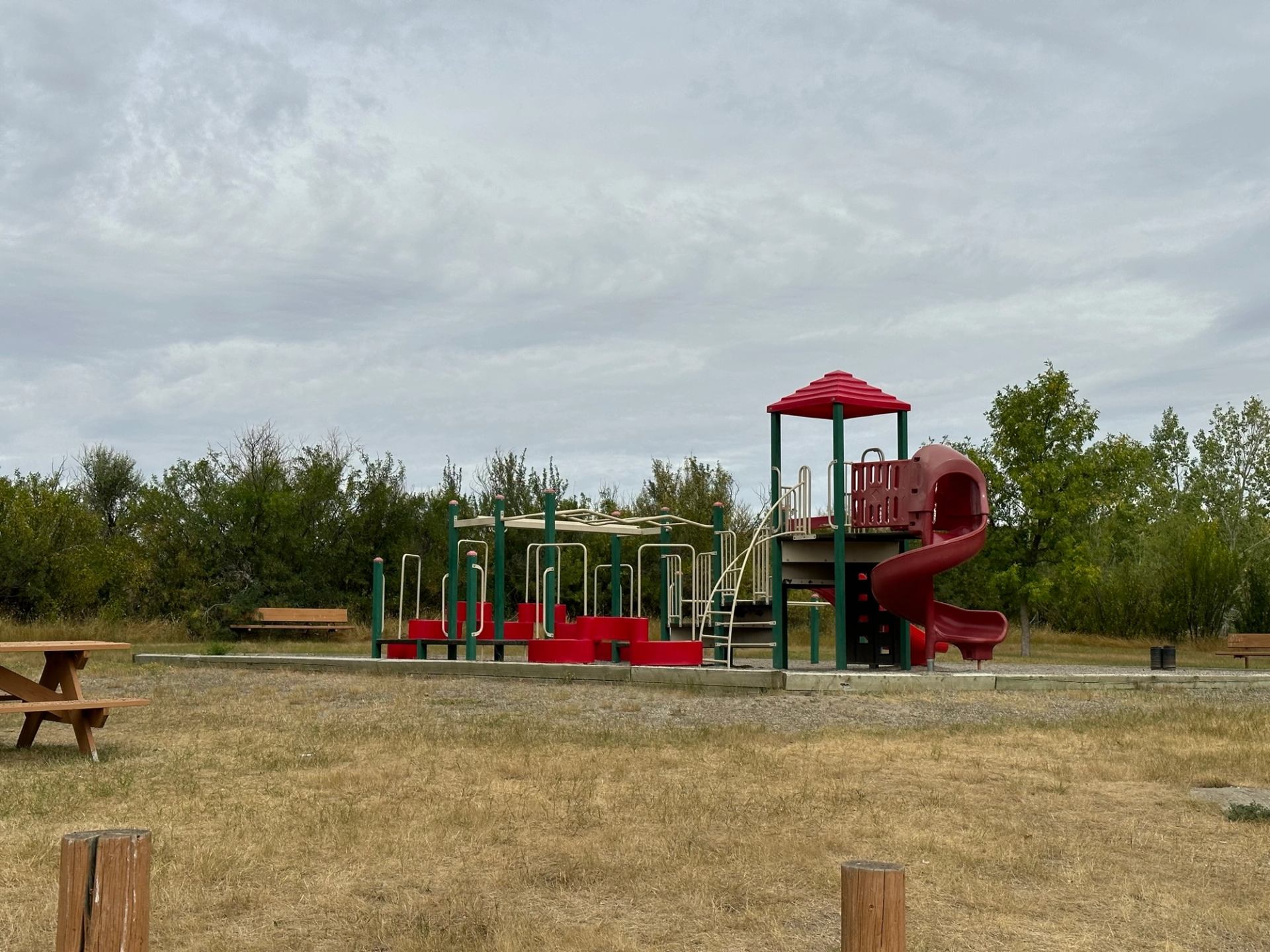 A colourful playground on dry grass with trees and benches nearby.