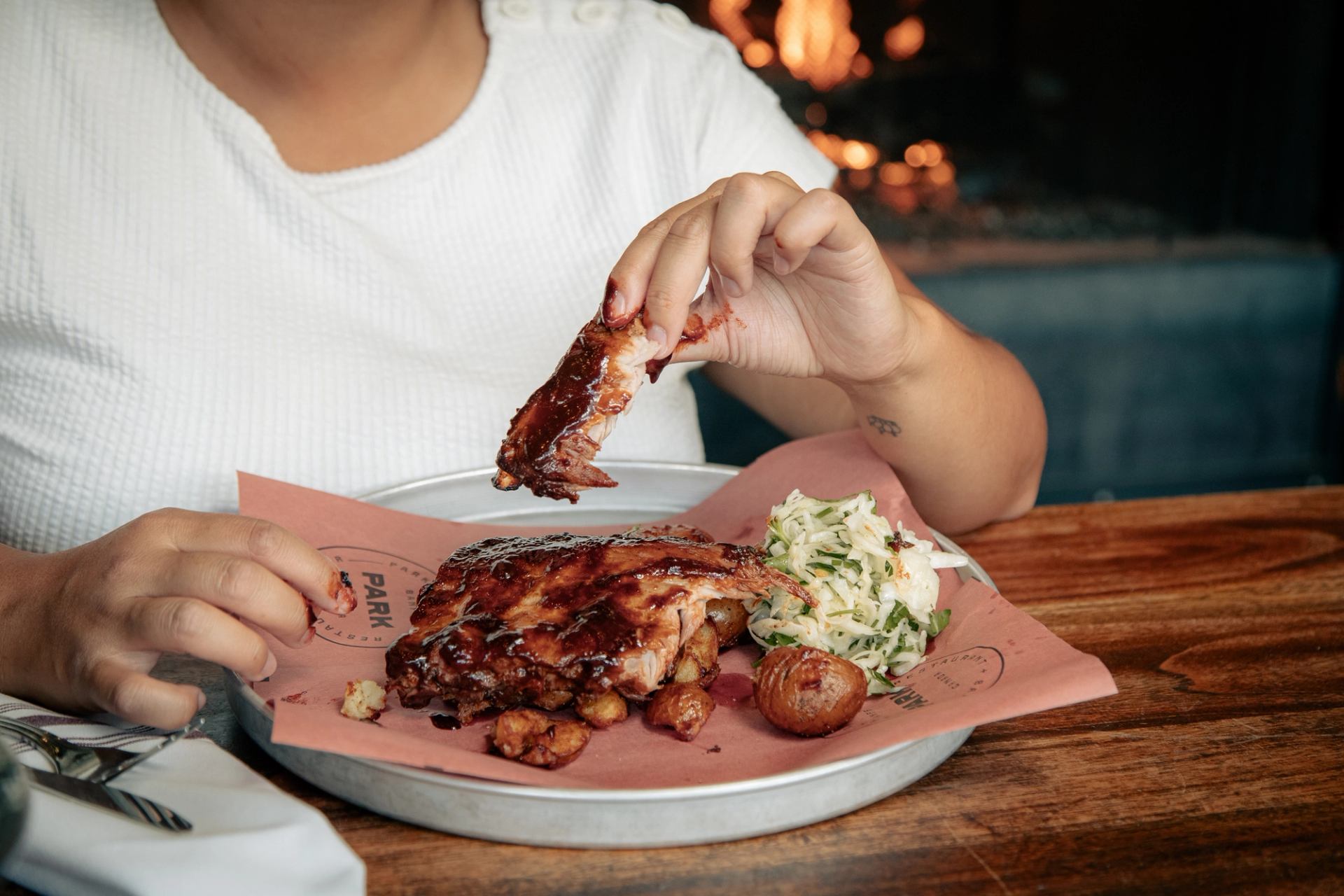A person holds a saucy BBQ rib from a platter with more ribs, coleslaw, and roasted potatoes.