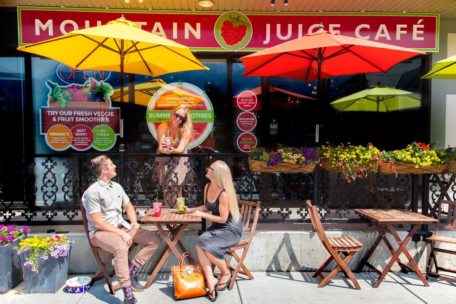 Outdoor seating at Mountain Juice Café with people, umbrellas, flowers, and bright signage.