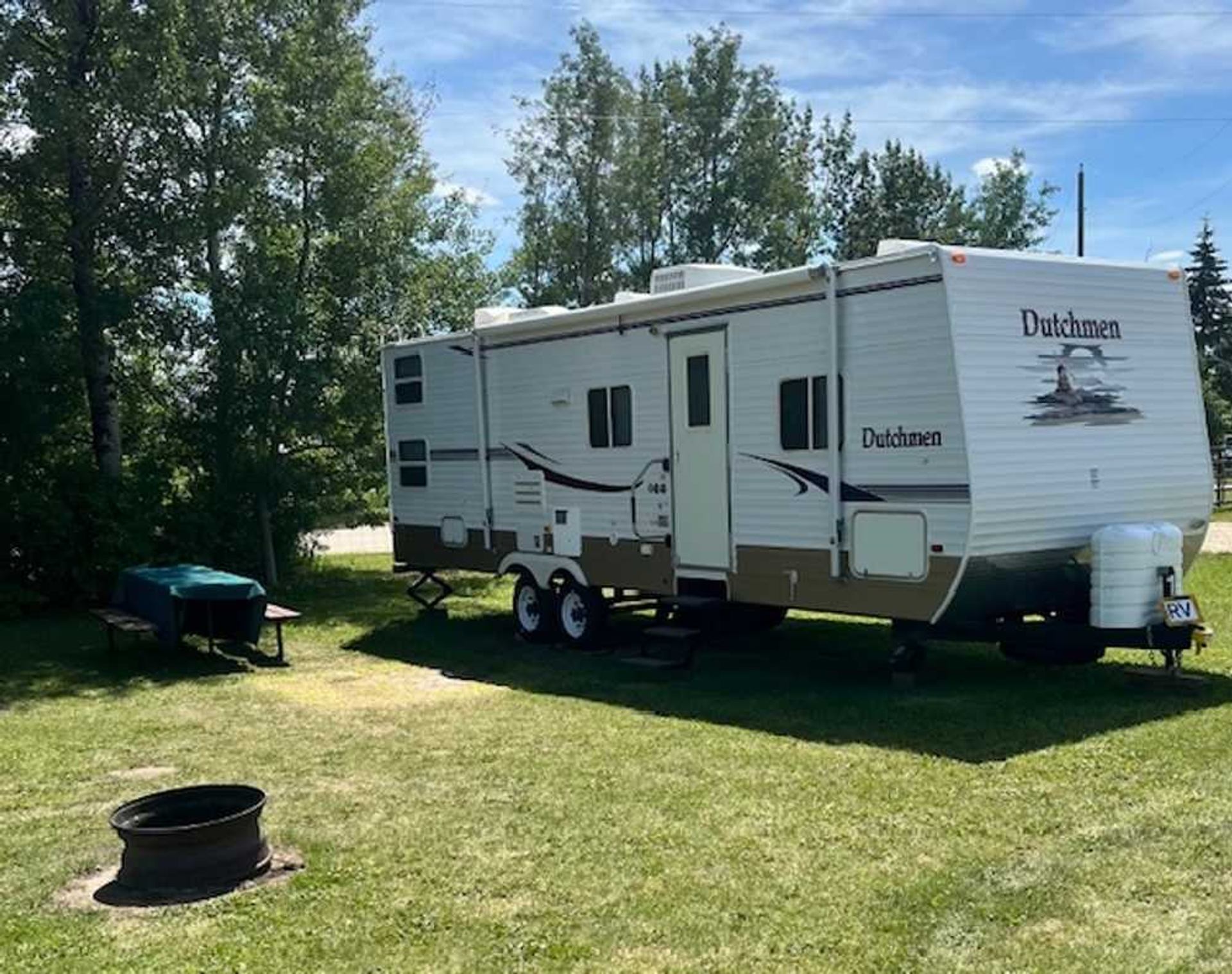 A white and brown Dutchmen travel trailer parked on a grassy campsite with a fire pit and picnic table.