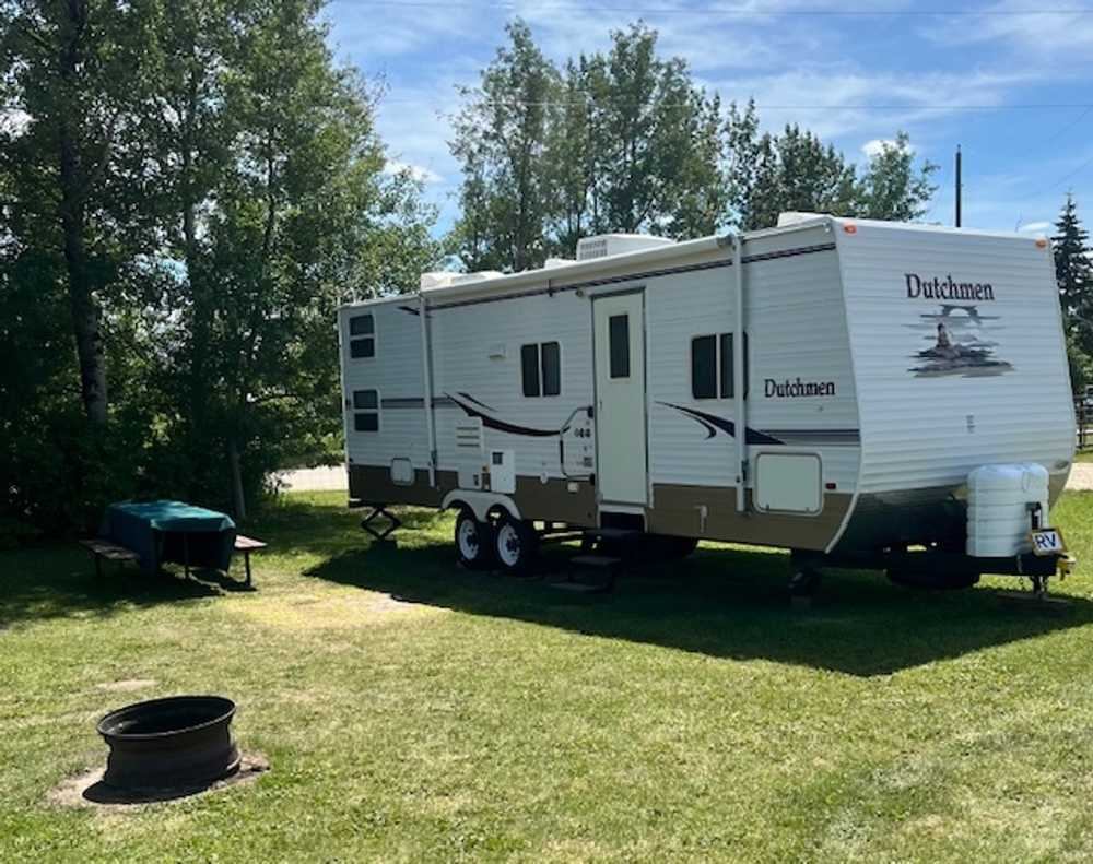 A white and brown Dutchmen travel trailer parked on a grassy campsite with a fire pit and picnic table.