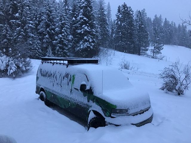 Green Cube Beyond Banff van covered in snow, parked amid snow-covered trees in a forested area.