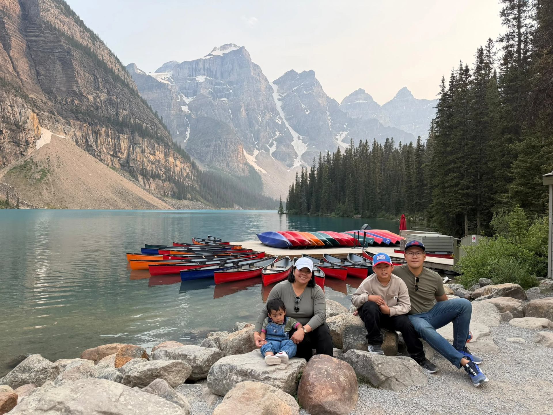 Four people sit on rocks beside canoes, a turquoise lake and rugged mountains.