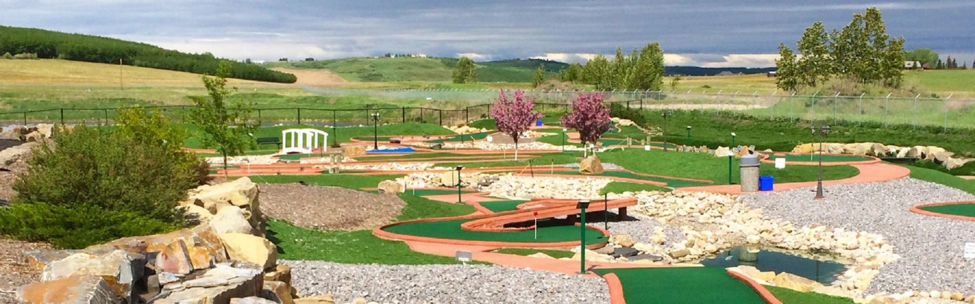 Mini-golf course with putting greens, pathways, rocks and small bridge, rolling hills beyond.
