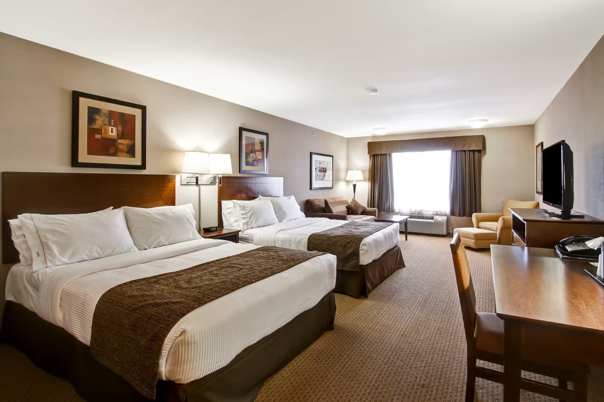Hotel room with two beds, sofa, armchairs, desk and television by window.