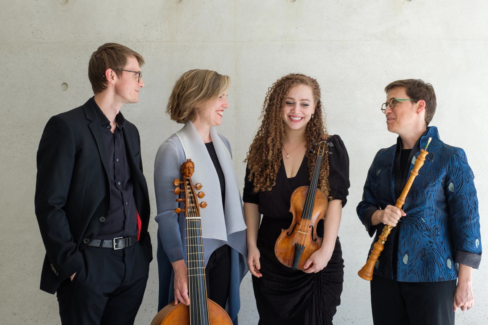 Four musicians stand against a light wall holding string and woodwind instruments.