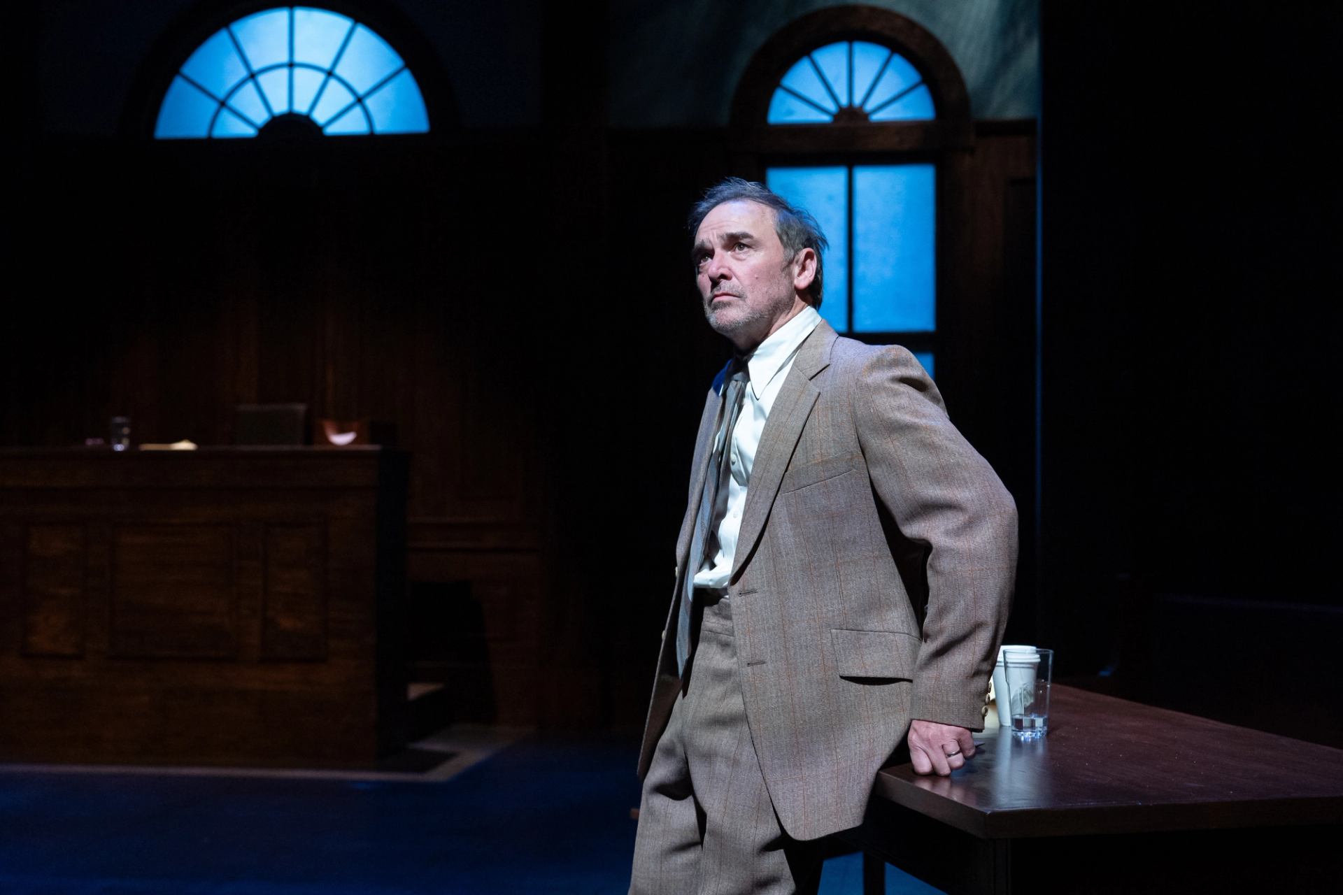 Actor in beige suit stands by courtroom table on dimly lit theatre stage.