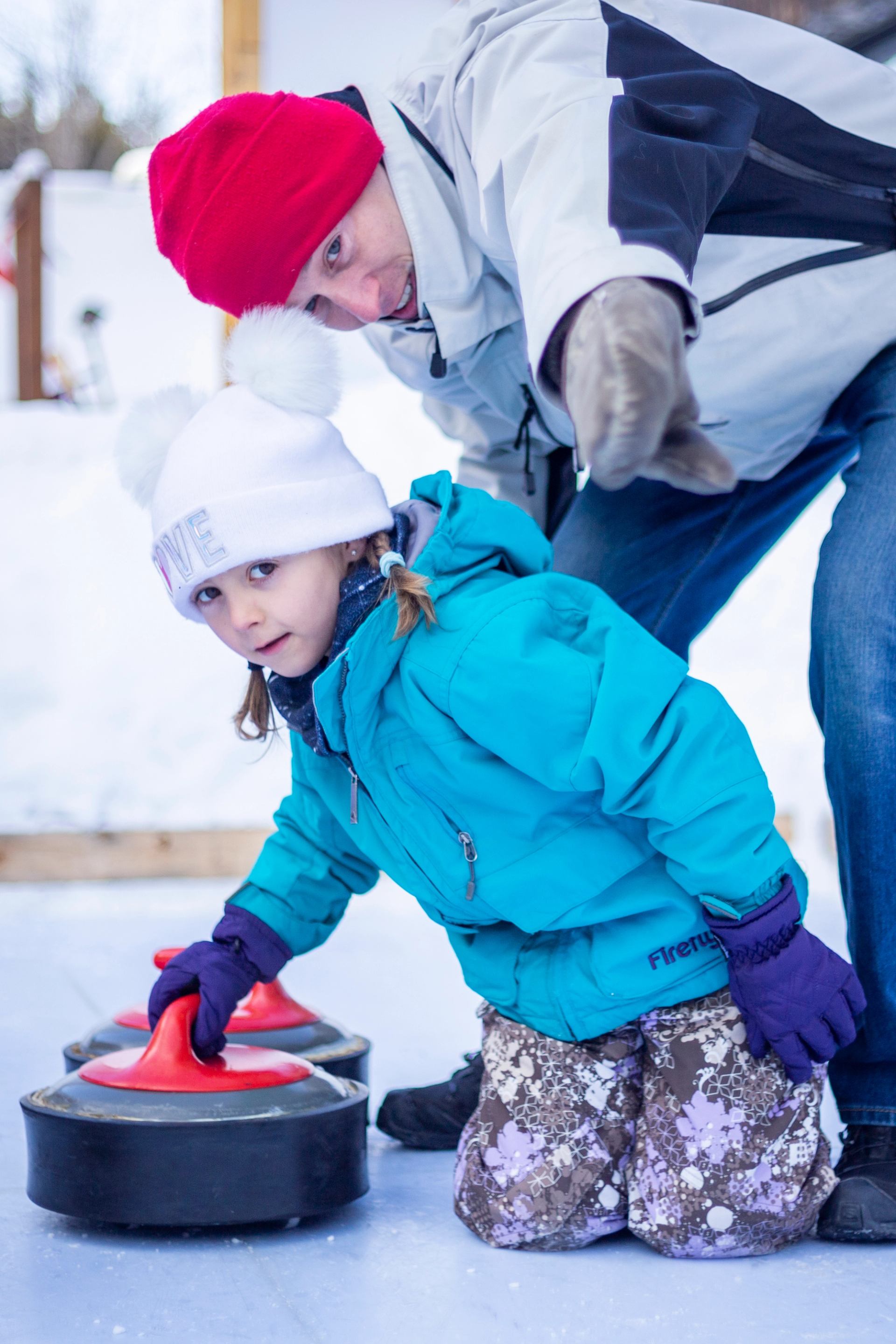 Child in winter clothing pushing a red curling stone on an outdoor ice sheet.