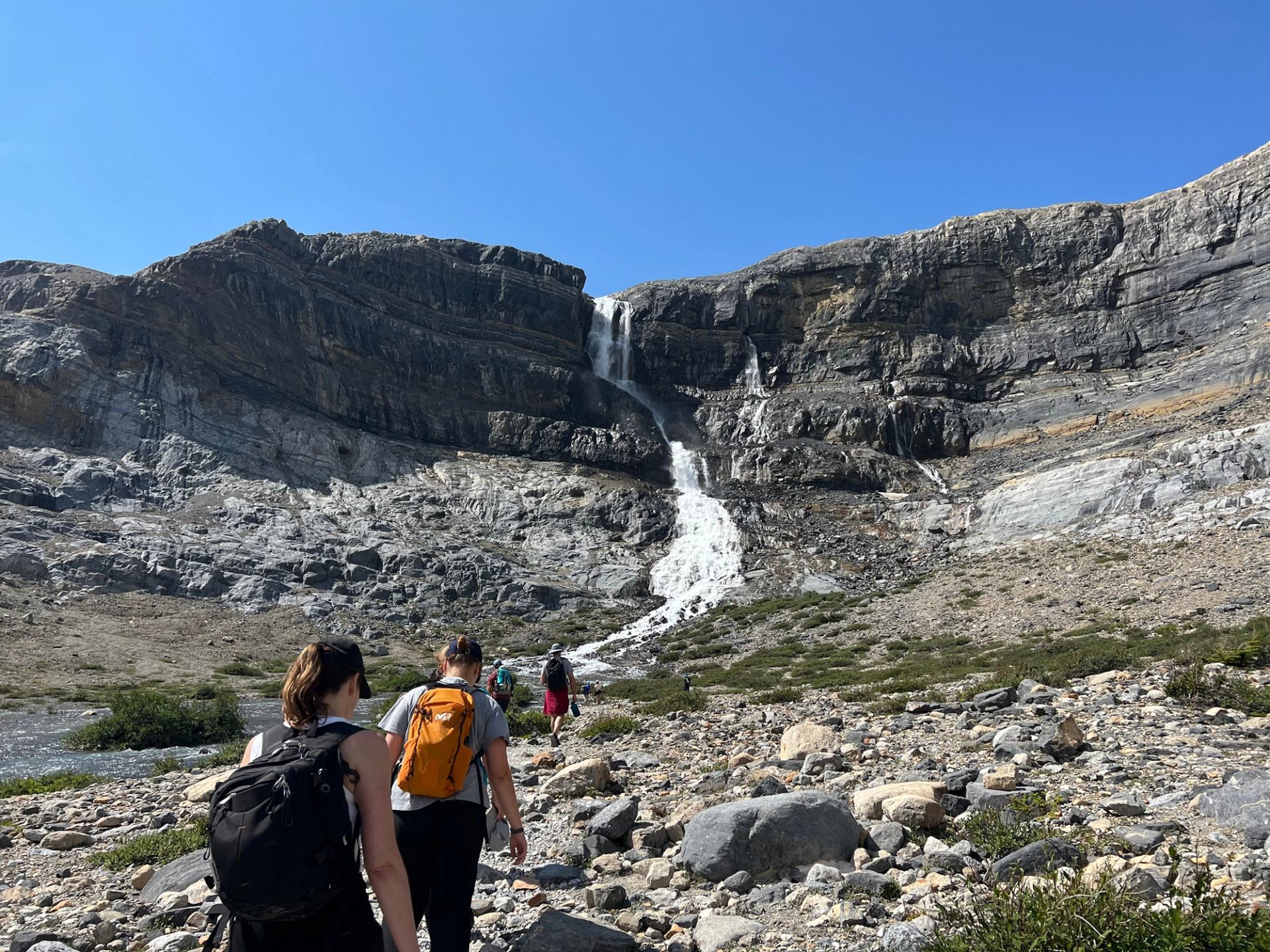 Hikers walking toward a cascading waterfall in a rocky alpine landscape under a clear blue sky.