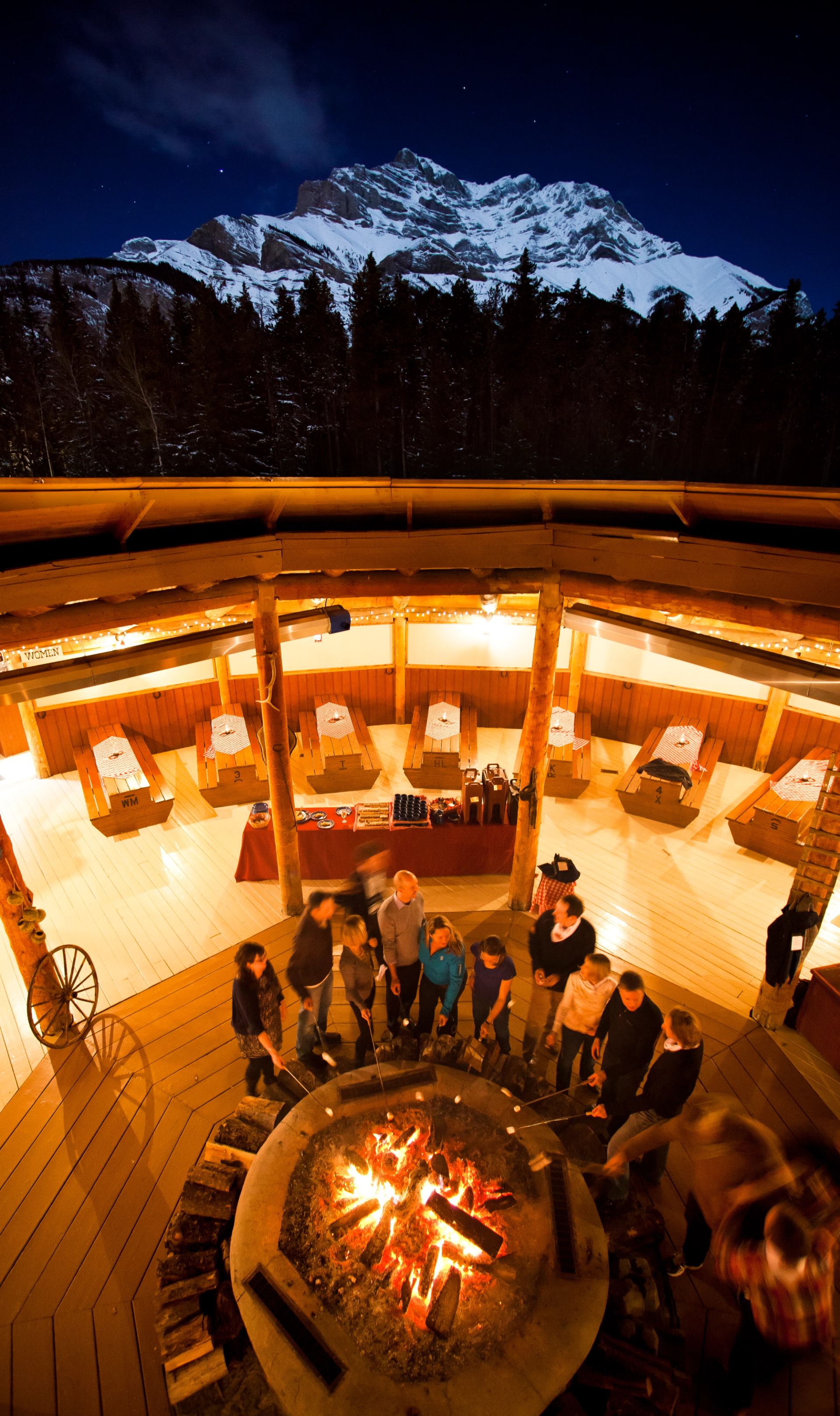 People roast marshmallows around a fire pit within a rustic wooden structure, with snow-capped mountains and a starry night sky visible above.