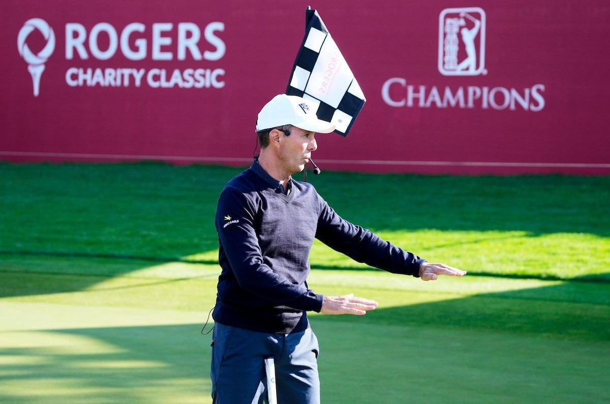 Golfer wearing a cap and headset, waving arms on a golf course before “Rogers Charity Classic” signs.