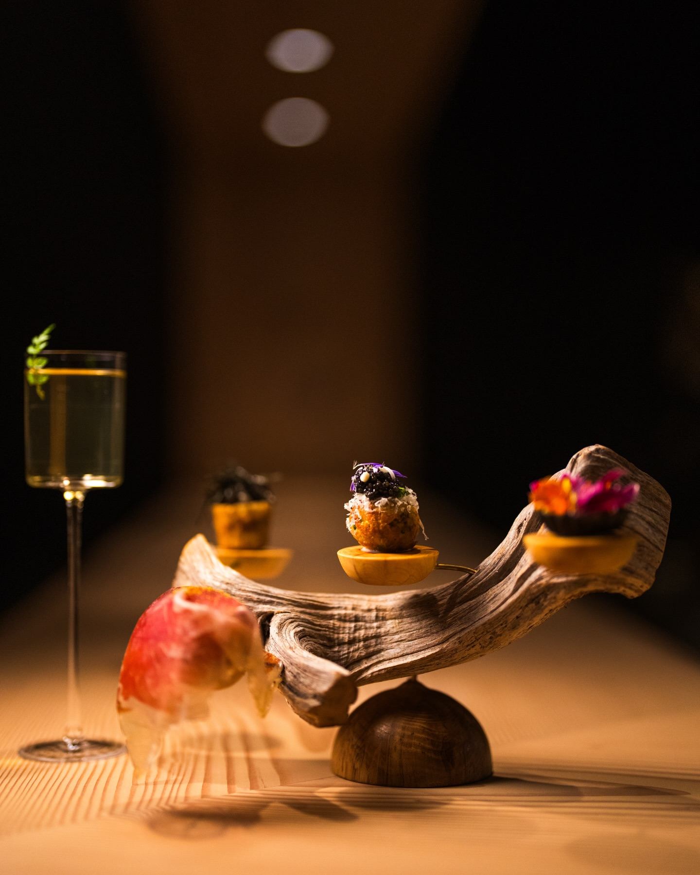 Gourmet bite-sized appetizers on sculpted wooden stand beside tall glass of sparkling drink.