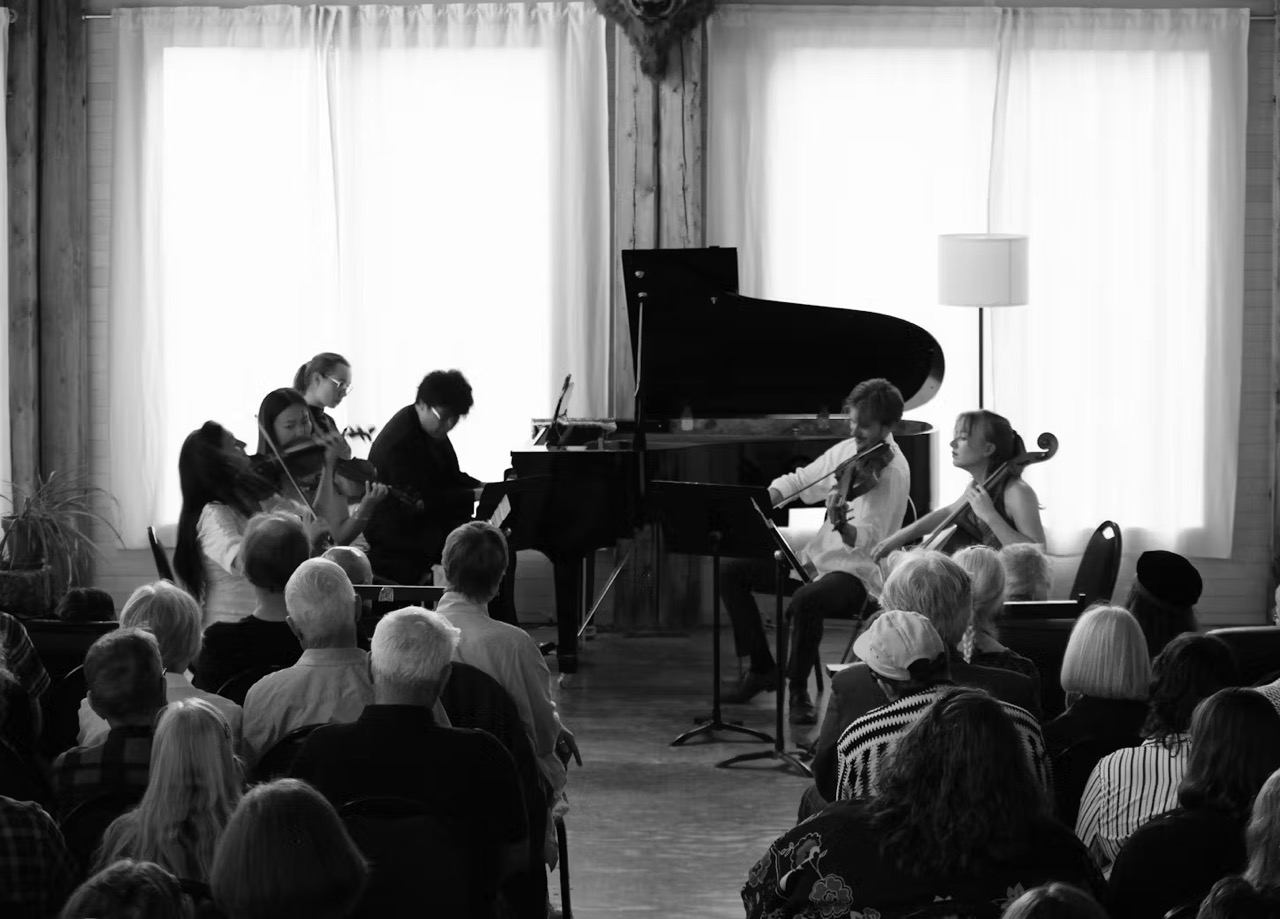 Audience seated as musicians perform string instruments beside a grand piano in a hall.