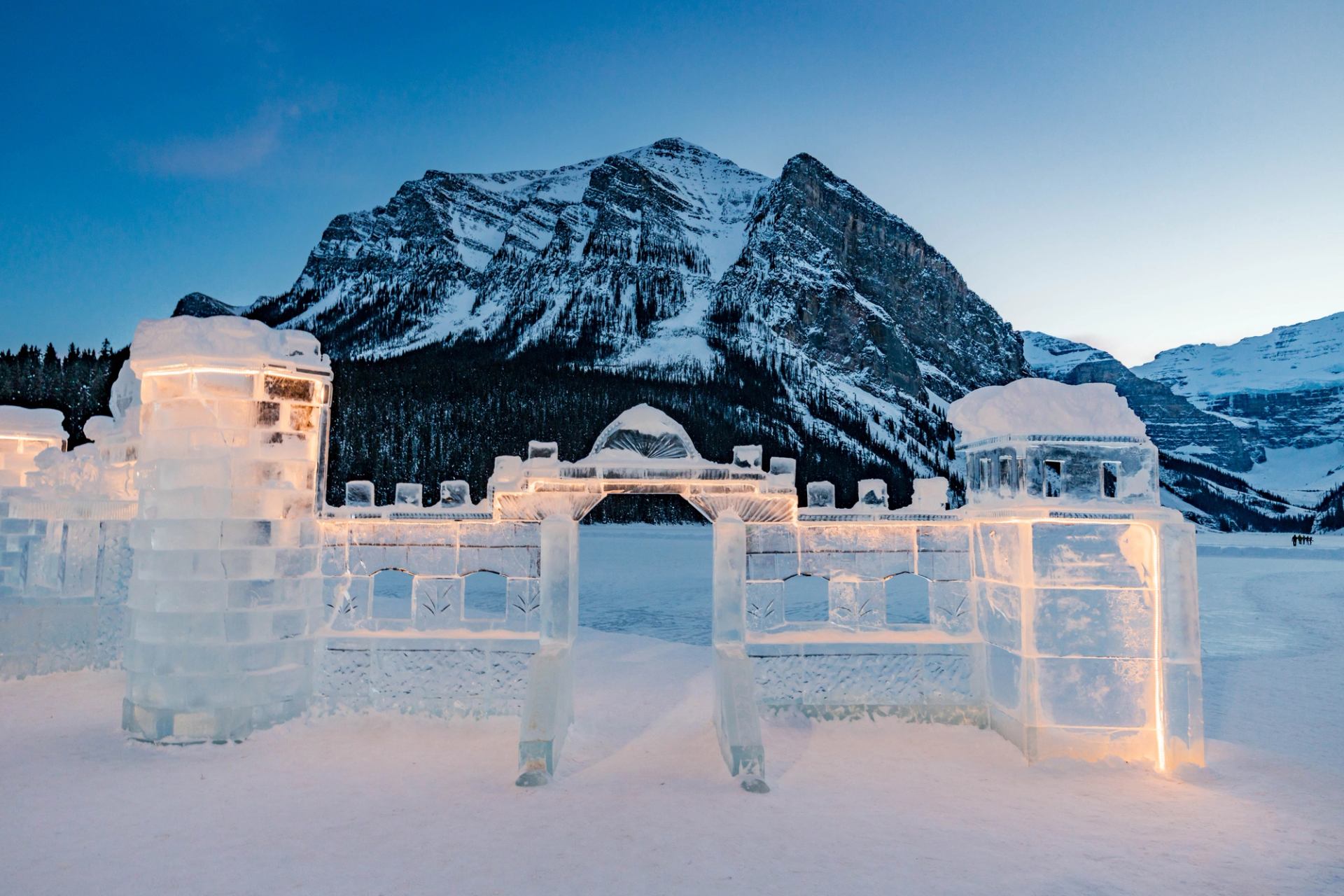 Illuminated ice castle with mountain backdrop at dusk in a snowy landscape.