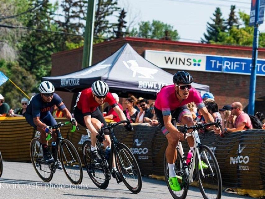 Cyclists race on a city street past spectators and a Bow Cycle event tent.