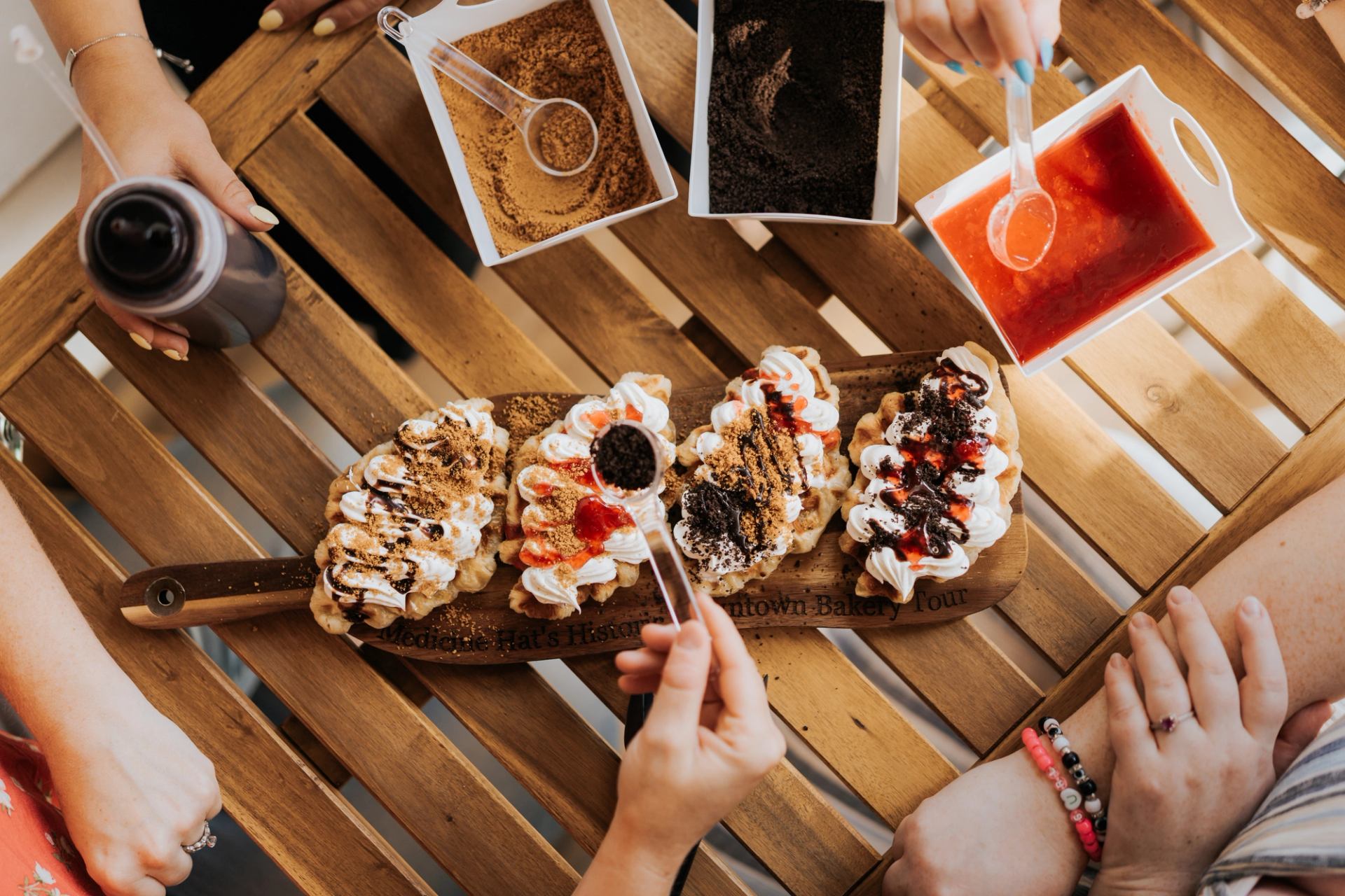 Hands topping assorted pastries with sauces and crumbs on a wooden table during a bakery tour.