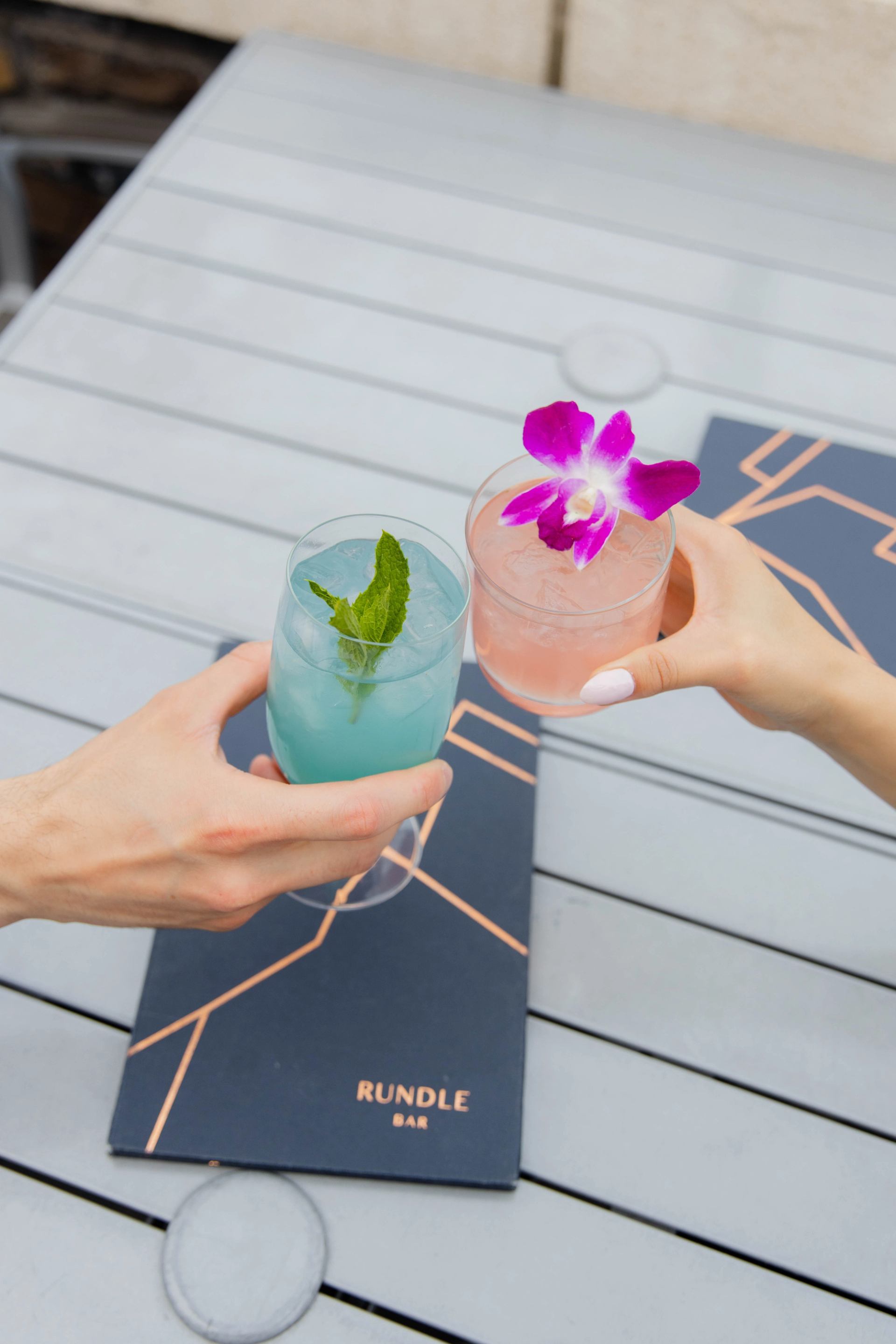 Two colorful cocktails on a gray table with Rundle-branded menus underneath.