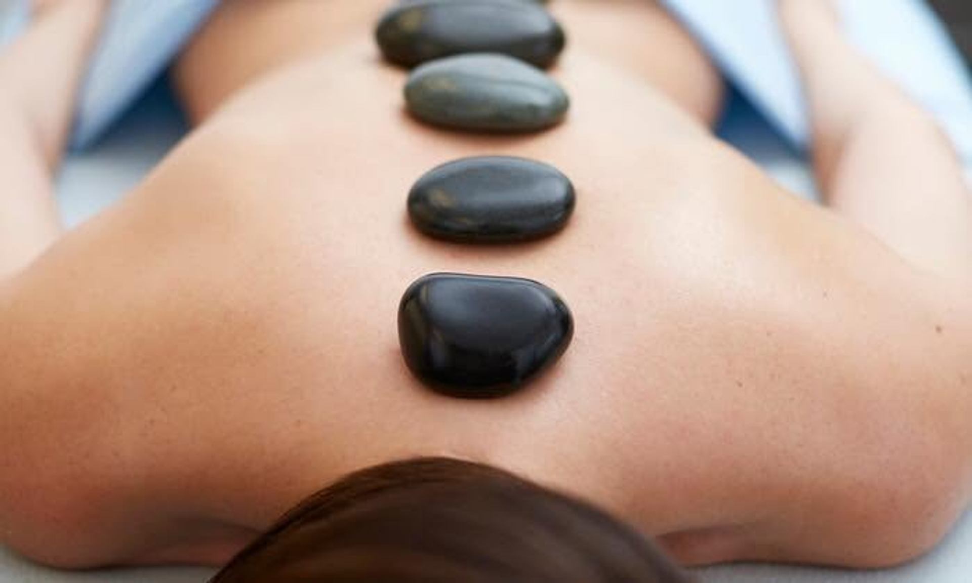 Person lying face down on massage table with smooth black stones along back.