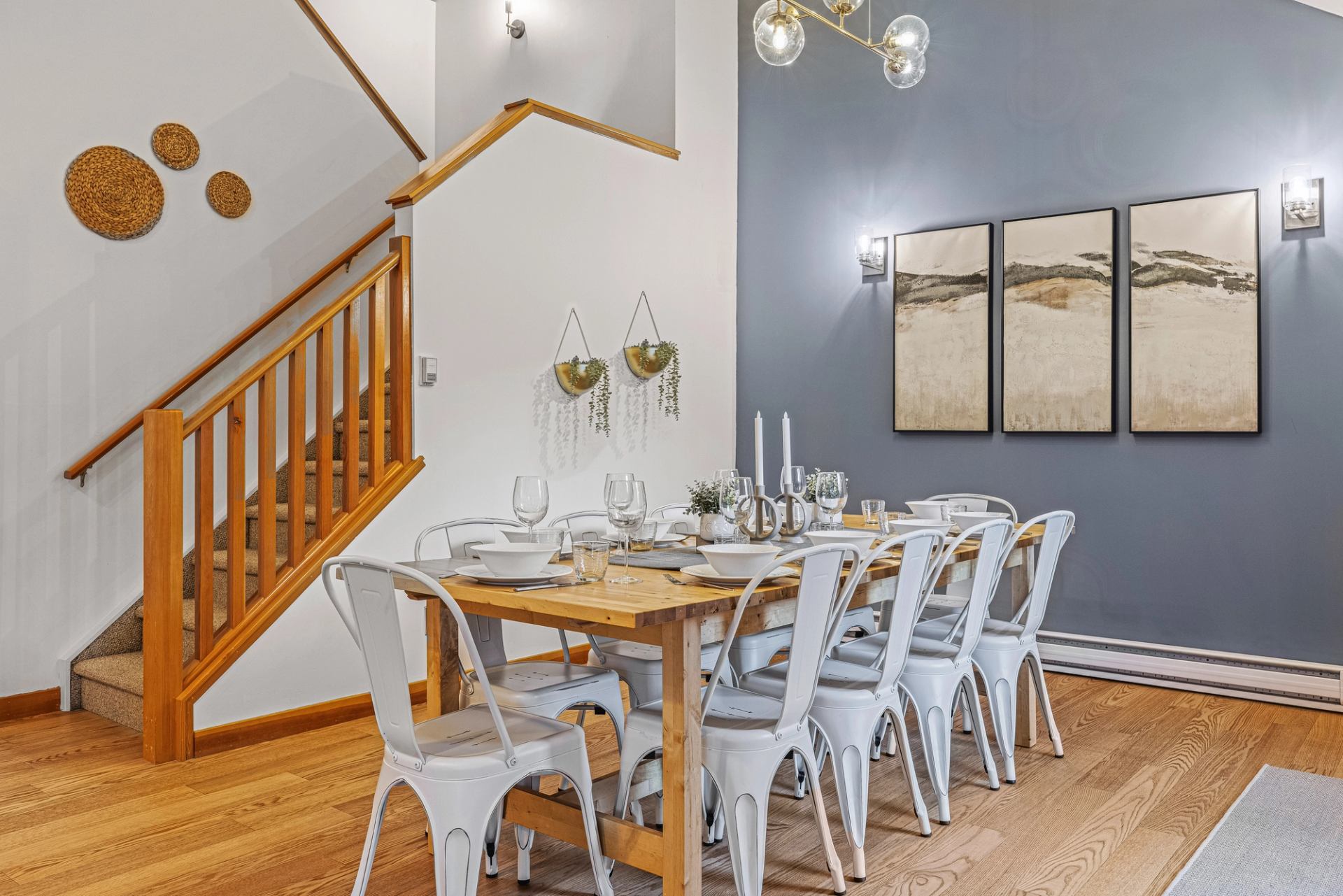 Dining room with long wooden table, white chairs, staircase and wall art.