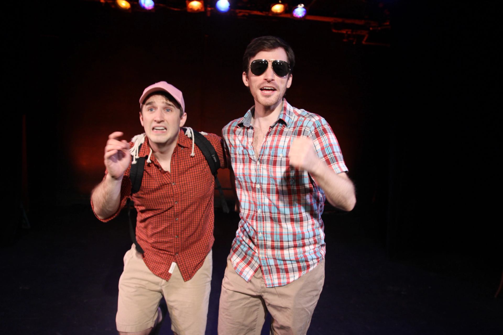 Two performers in plaid shirts onstage, one wearing sunglasses, under colourful stage lights.