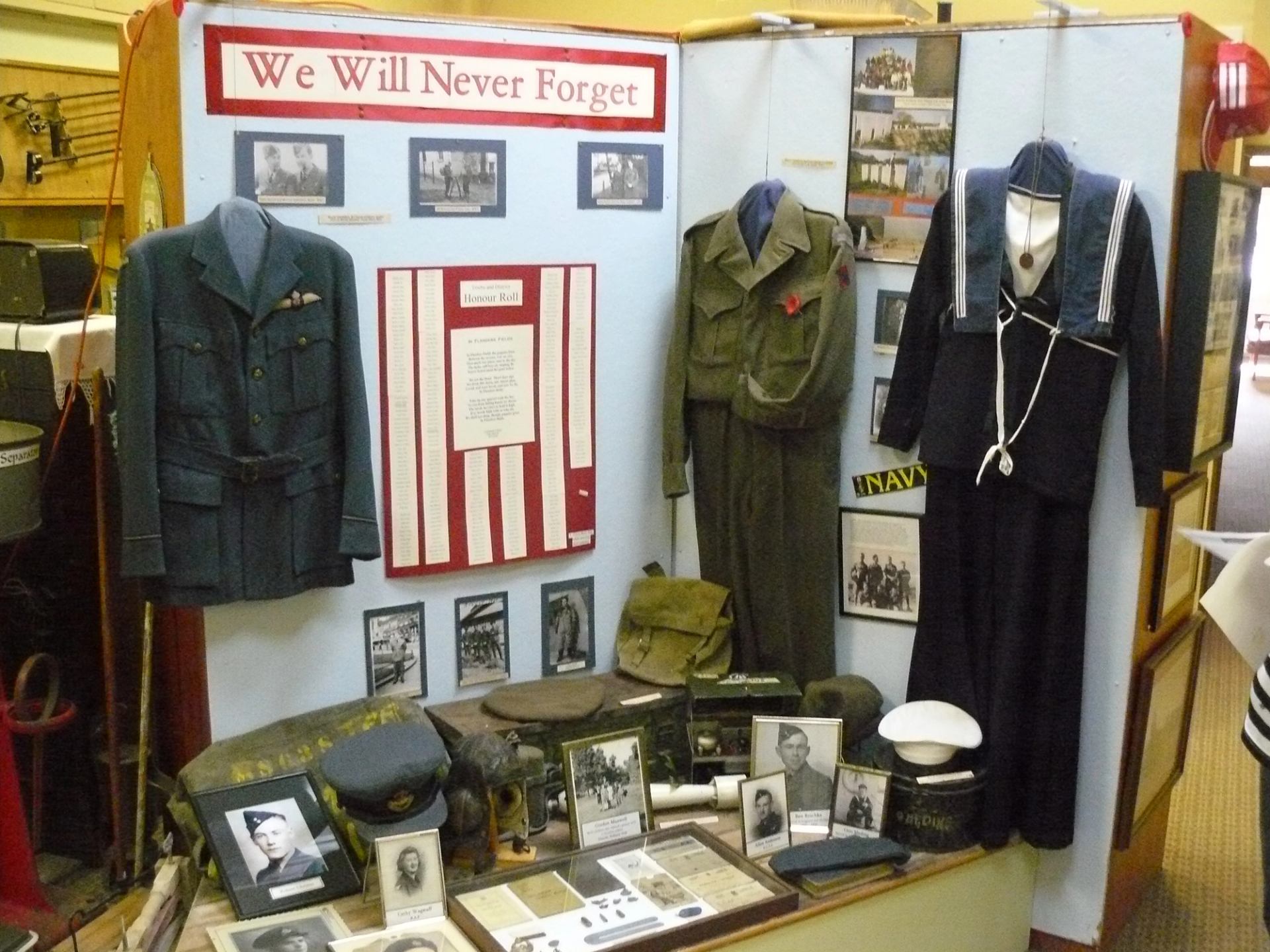 Display of vintage military uniforms, hats, photos, and gear under a “We Will Never Forget” sign