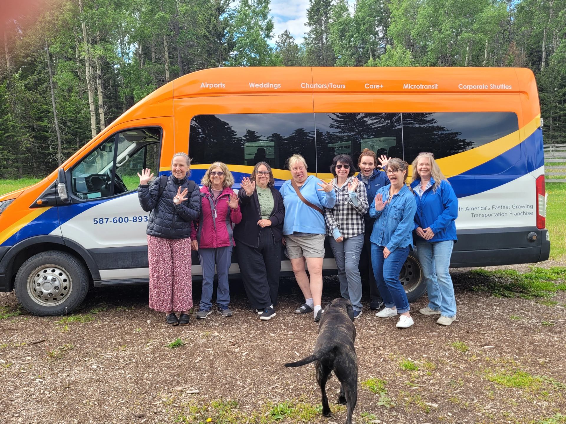 A groups of travelers standing in front of a shuttle van, with a friendly dog approaching