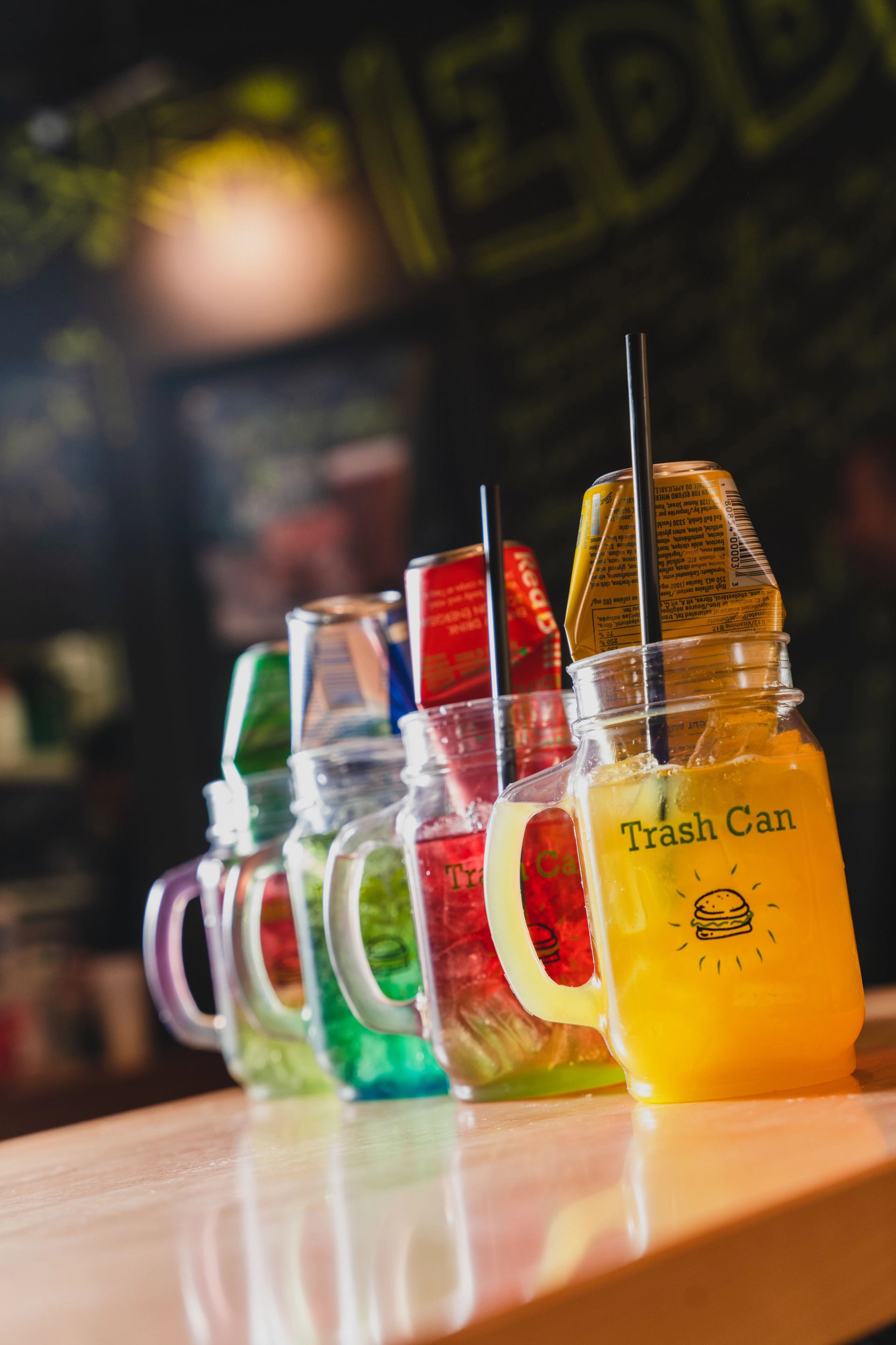 A row of five colorful mason jar drinks, each with an inverted beverage can/carton, on a wooden counter. 