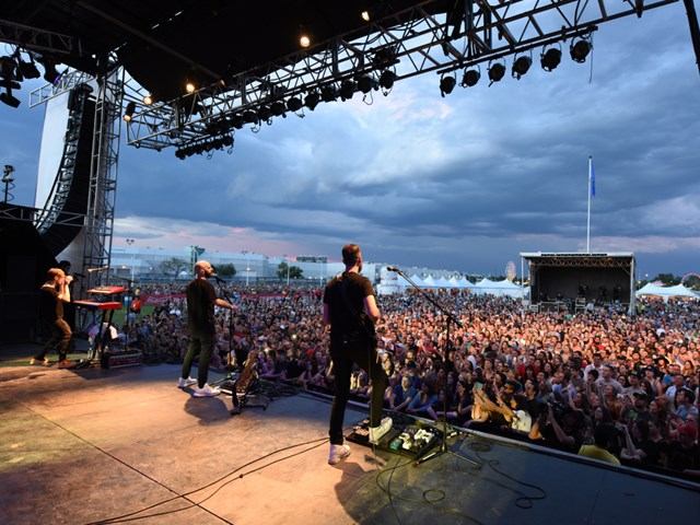 Band plays on outdoor stage with crowd and cloudy sky at K Days festival.