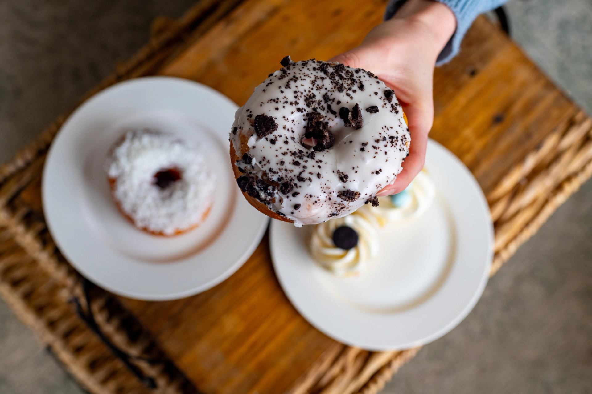 Hand holding a cream‑topped donut above plates of pastries on a wooden table.
