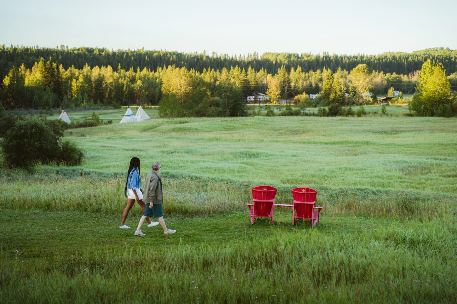 Two people walk past red lawn chairs in a wide grassy field with distant tipis.