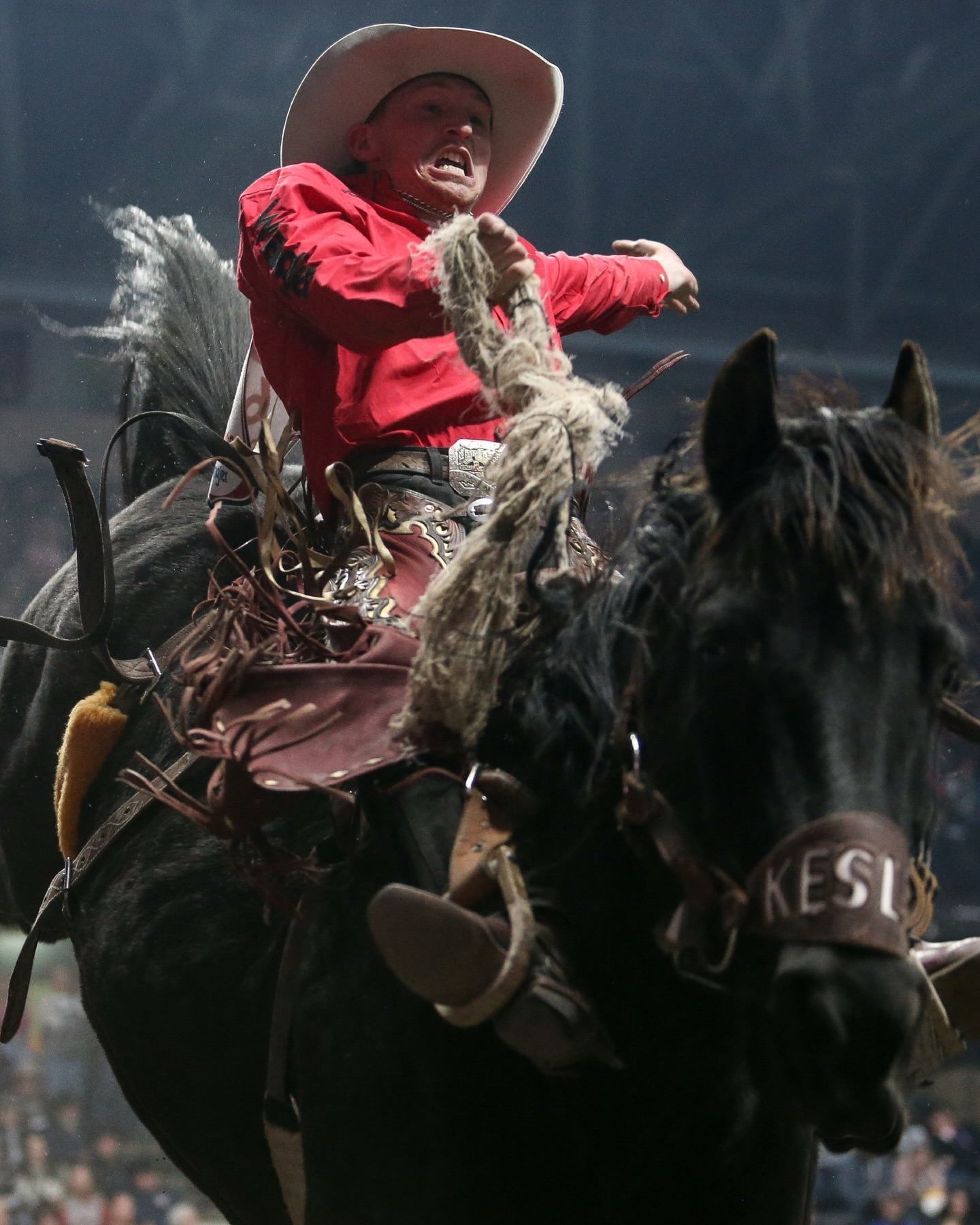 Rodeo rider in red shirt grips rope while riding bucking horse in arena.