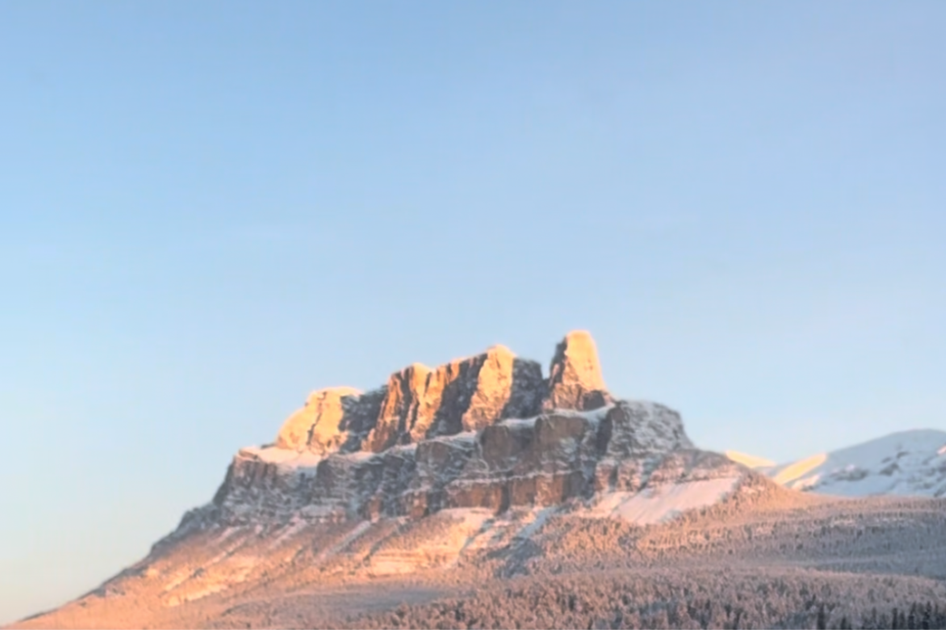 Sunlit rocky peak dusted with snow under a clear pastel sky.