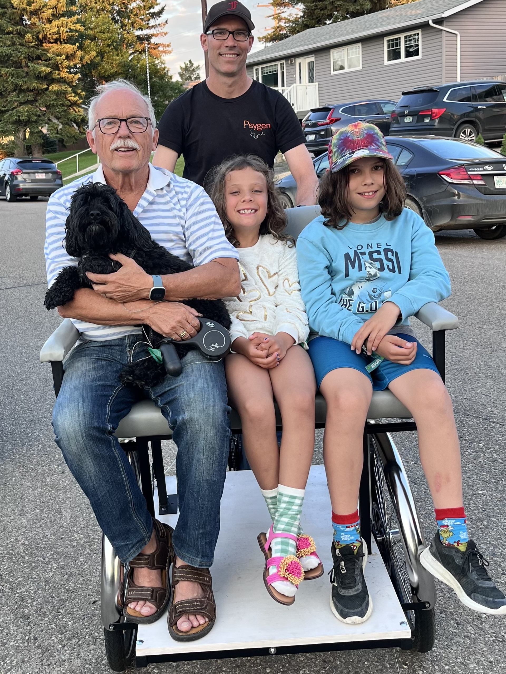 Three seated people and a standing man pose with a black dog outdoors.