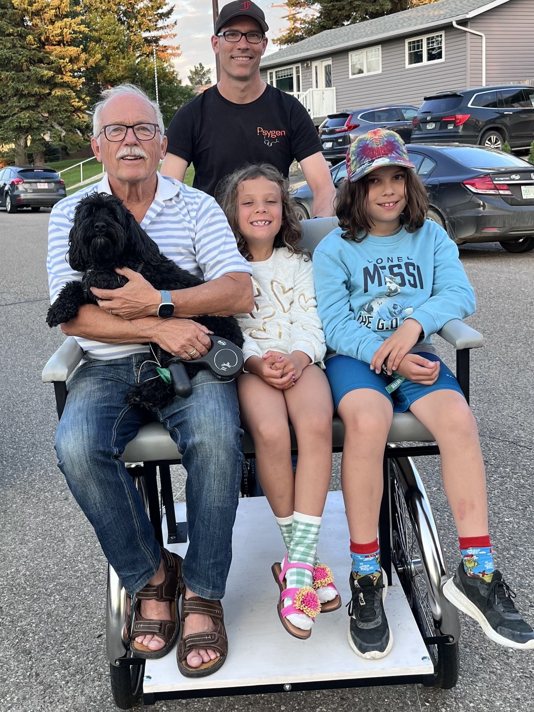 Three seated people and a standing man pose with a black dog outdoors.