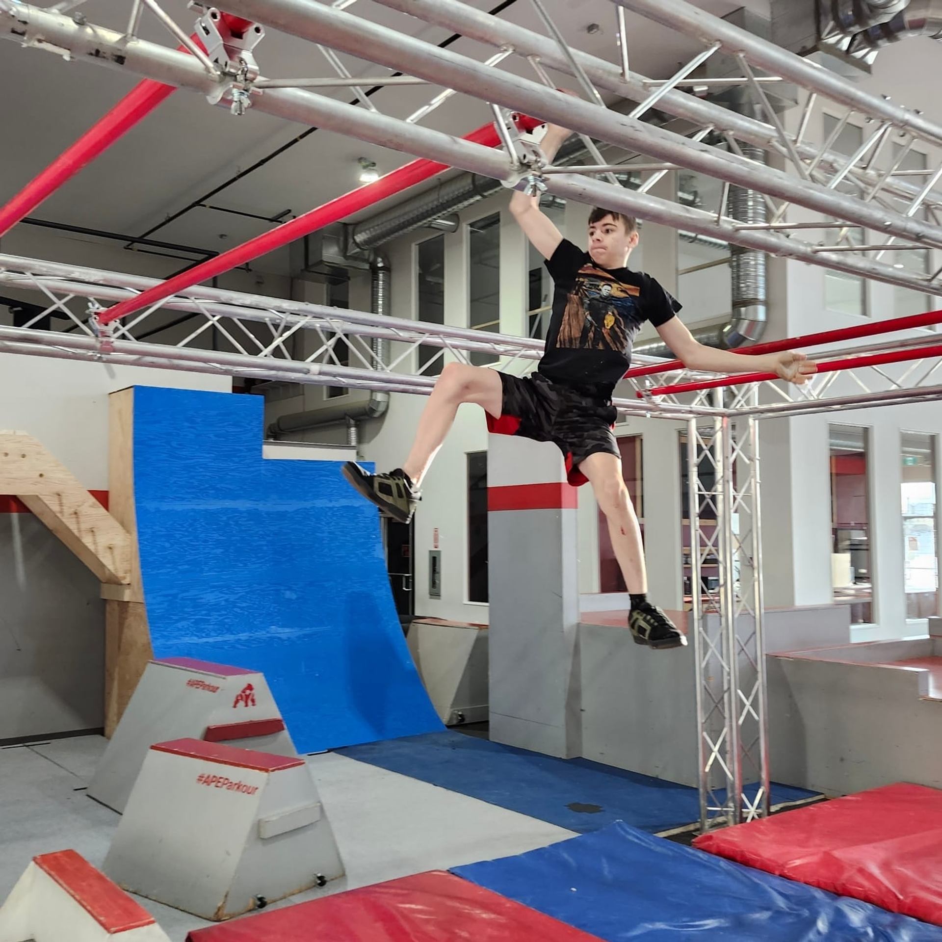 A young man hangs from an overhead bar on an indoor obstacle course.
